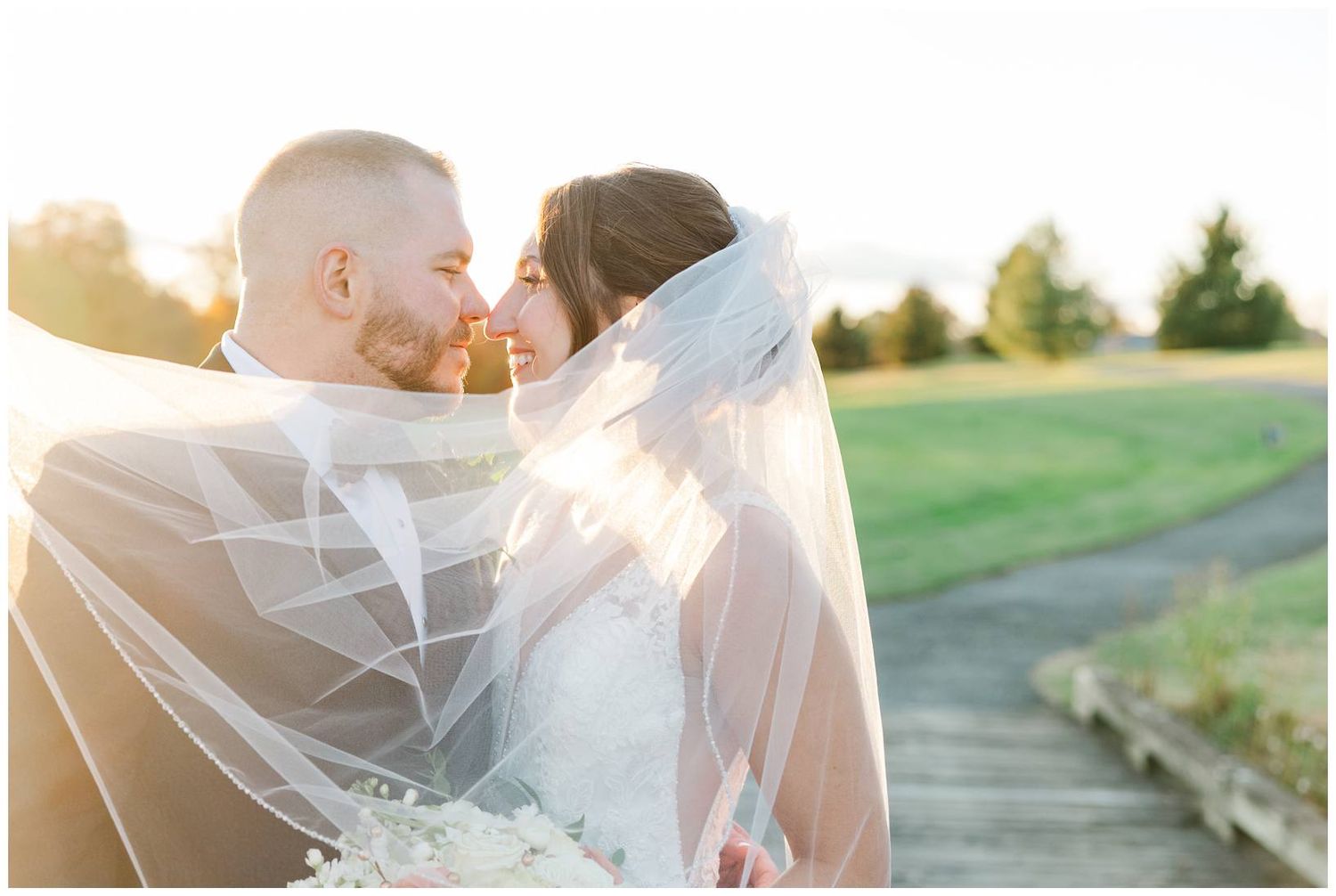 Romantic sunset portrait of newlyweds embracing under flowing veil on golf course path.