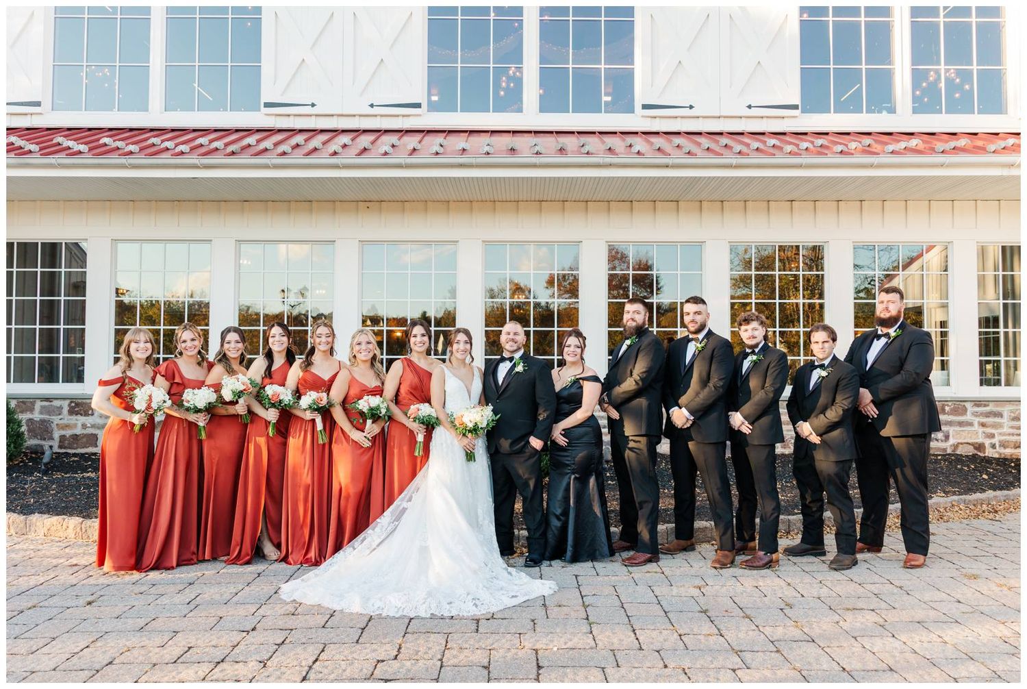 Elegant wedding party lined up outside white venue building with stone facade and large windows.