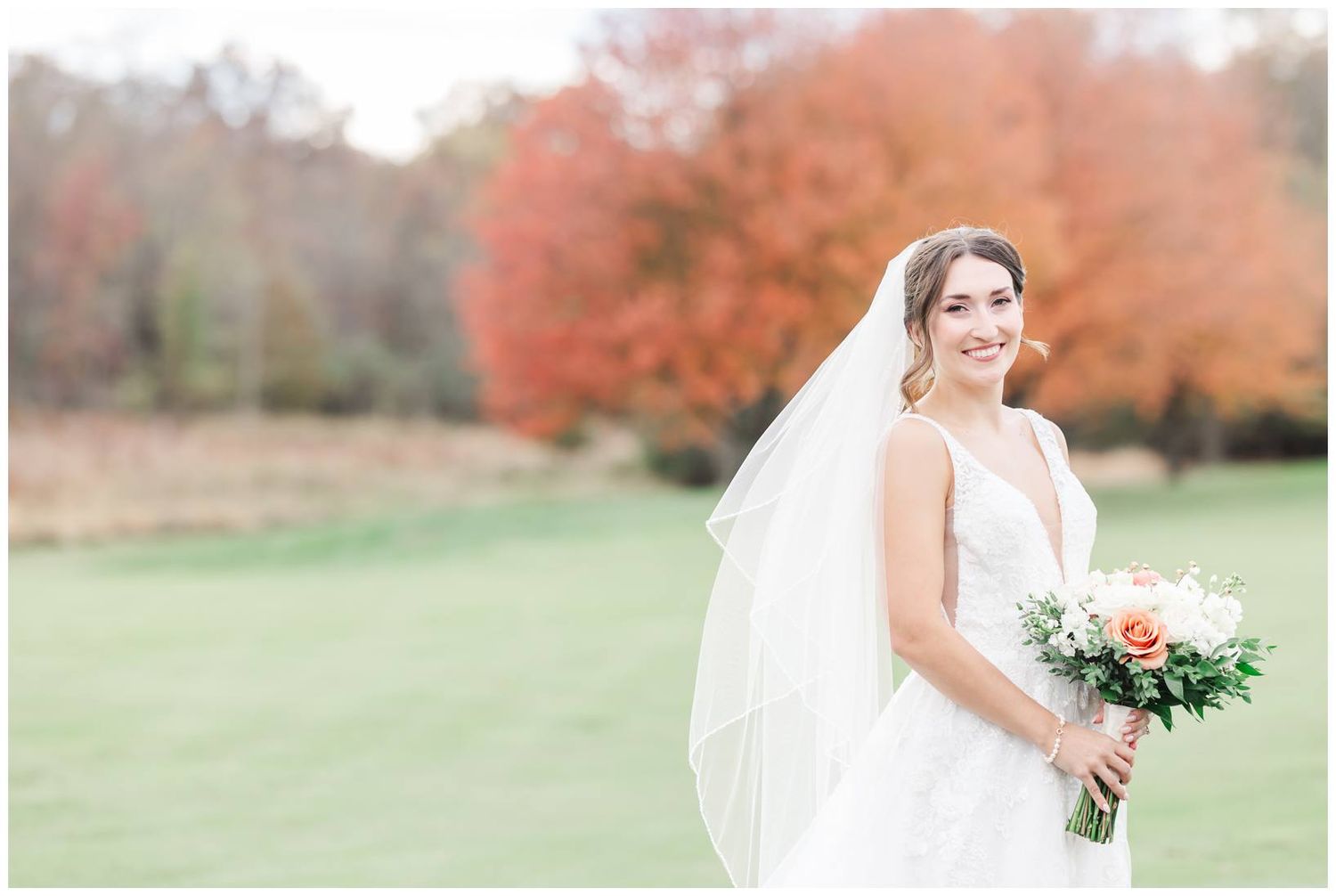 Bride in white dress holds bouquet while standing on golf course with fall colors in background.