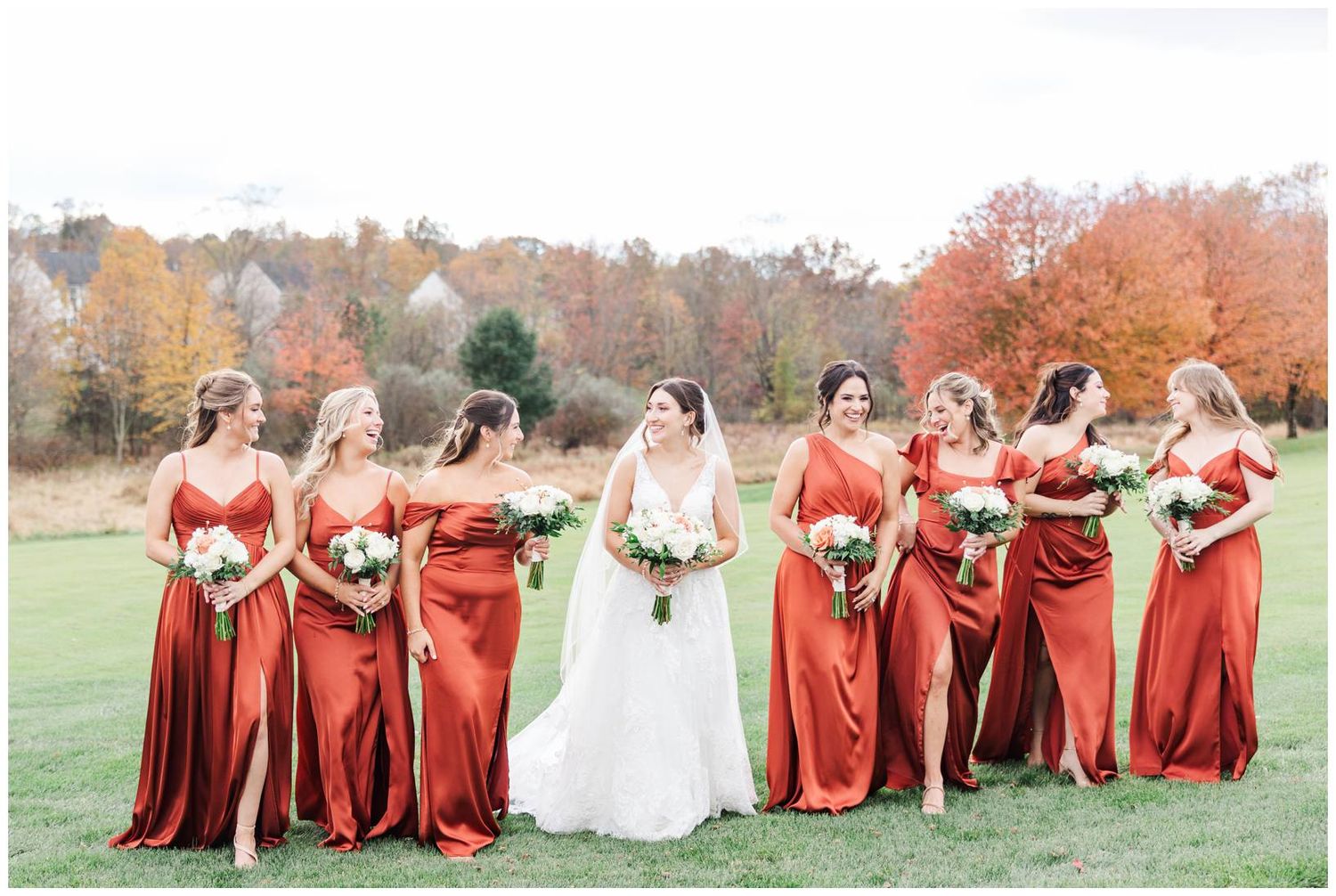 Wedding party poses outdoors with bridesmaids wearing elegant rust-colored dresses against autumn foliage.