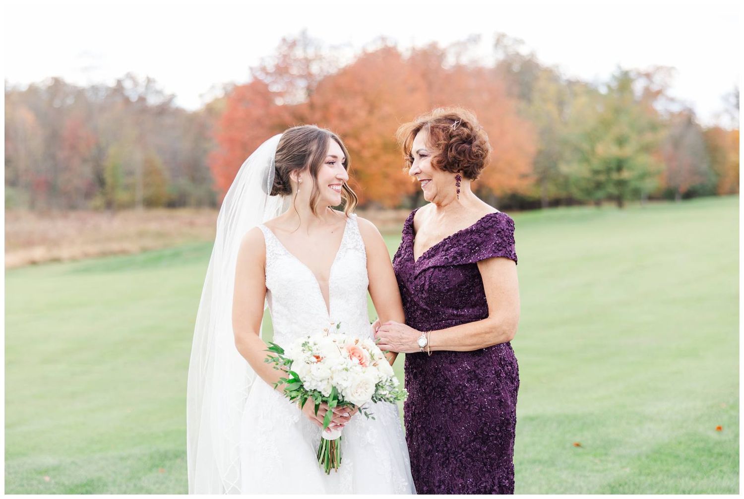 Wedding party members in white and purple dresses pose together on grassy field during fall season.