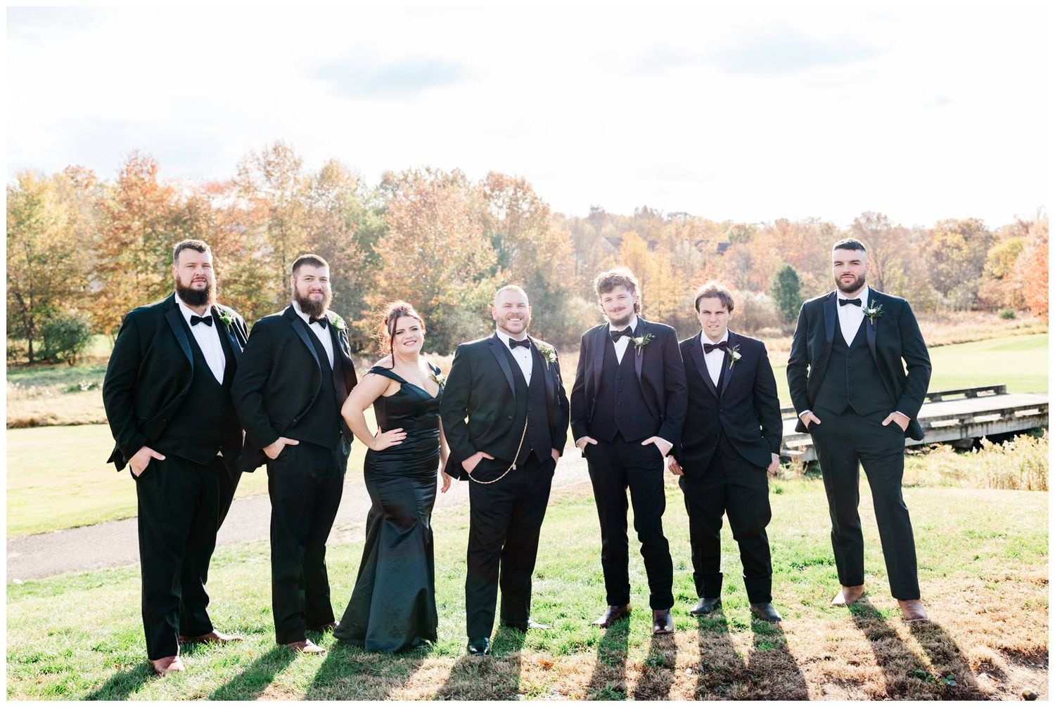 Wedding party poses together on grassy hill during autumn sunset with fall foliage in background.