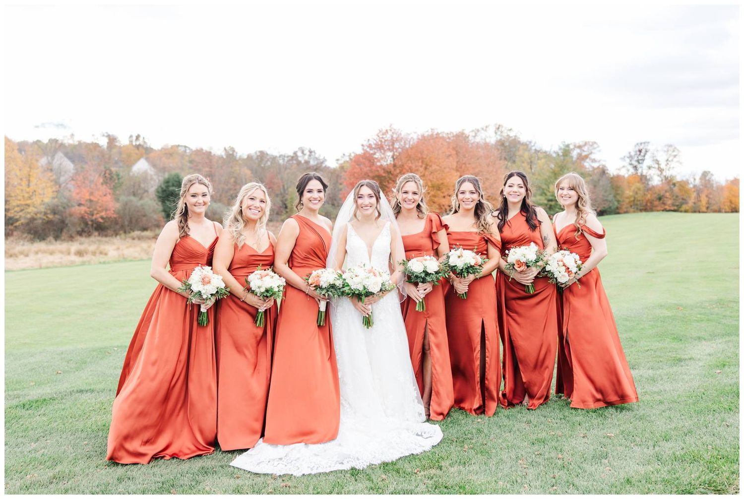 Bridesmaids in matching coral-colored dresses stand together on a grassy hillside during fall.