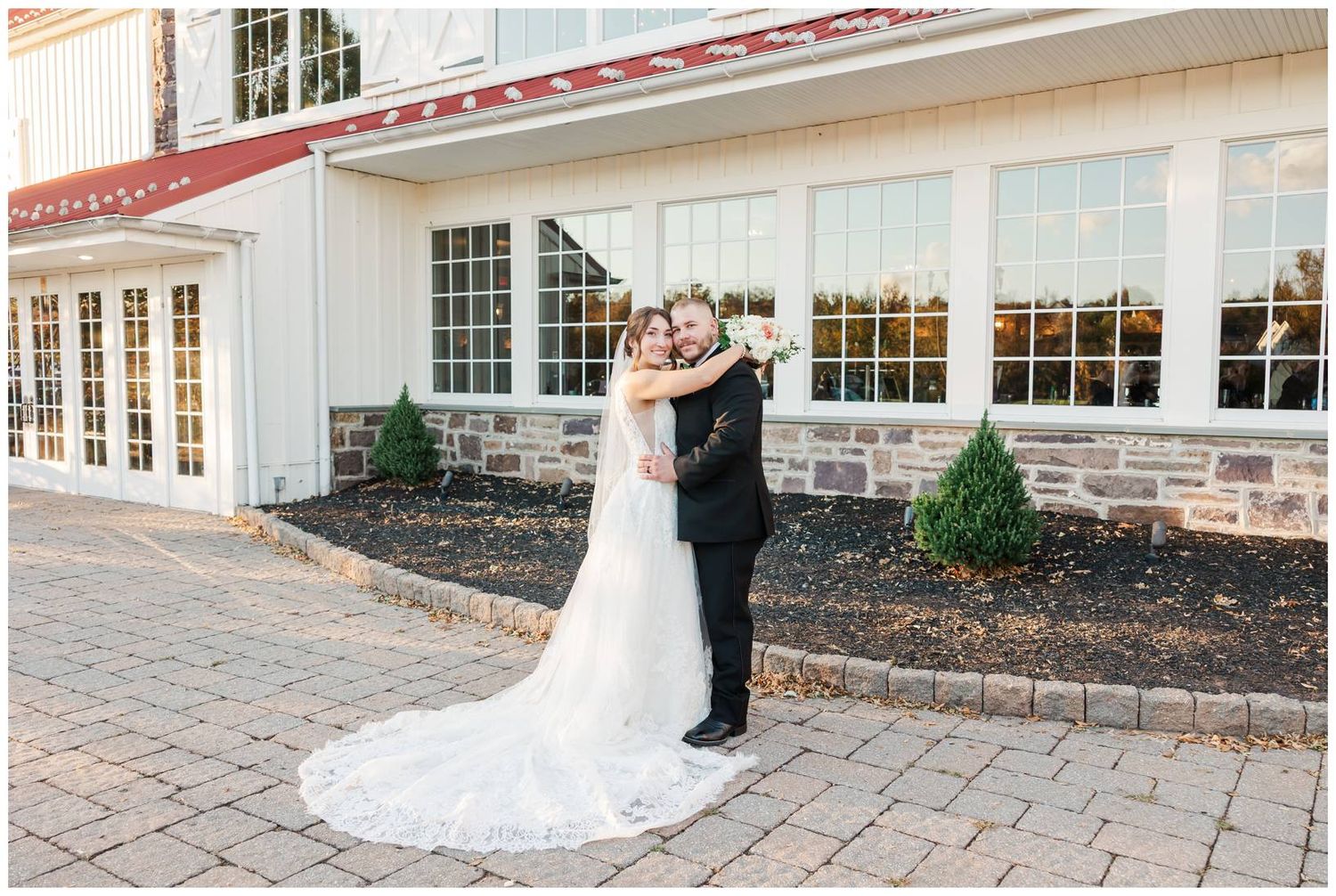 A couple stands together on a stone paved courtyard outside a white building with many windows.