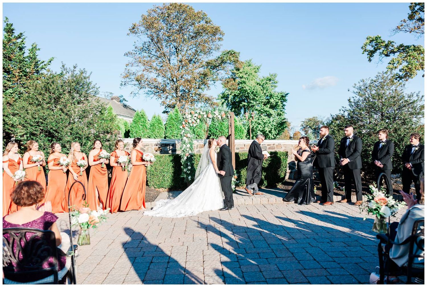 Outdoor wedding ceremony with bridal party in coral dresses and guests seated on lawn.