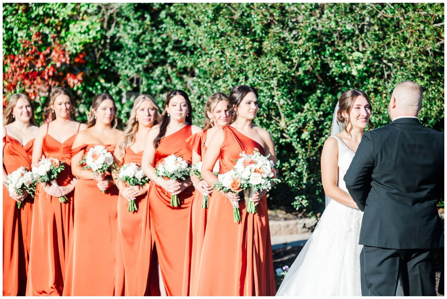 Wedding party lineup outdoors with coral bridesmaid dresses against a green hedge backdrop.