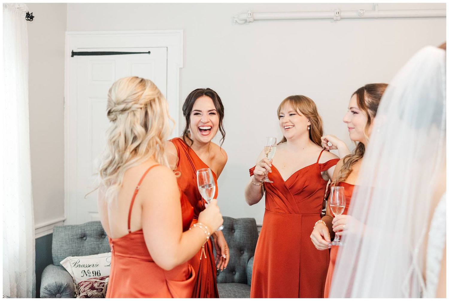 Bridesmaids in coral-colored dresses share laughter and champagne during wedding day preparations.