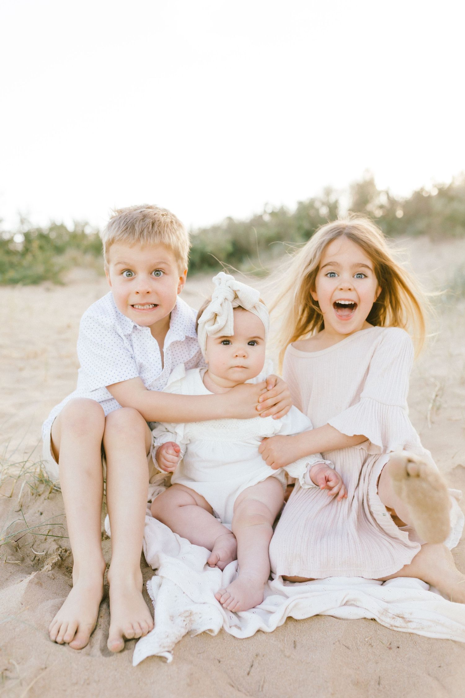 Children wearing white outfits sit together on a sandy beach during a bright summer day.