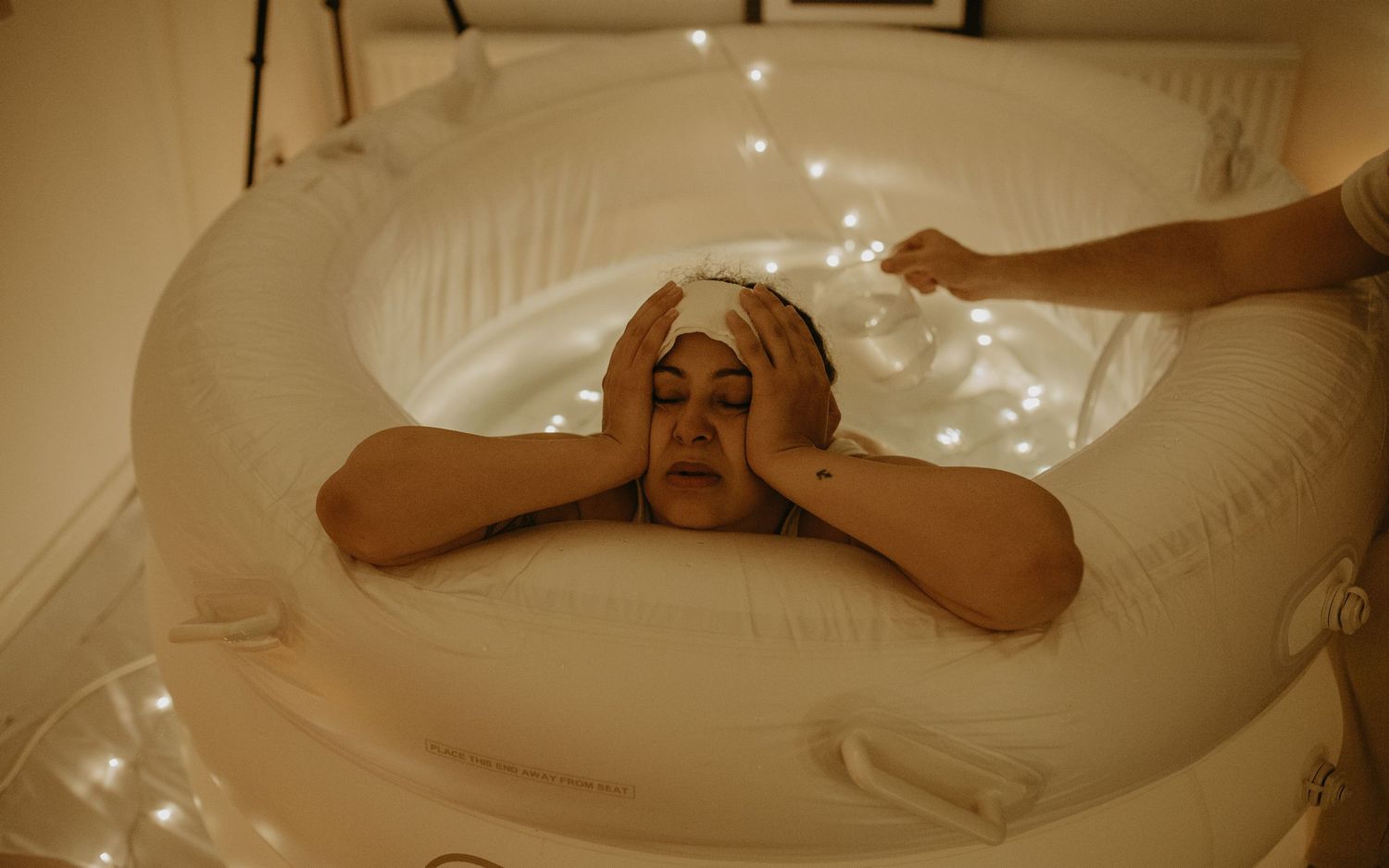 Person relaxing in white bathtub with hands covering their face in warm atmospheric lighting, birth photographer london