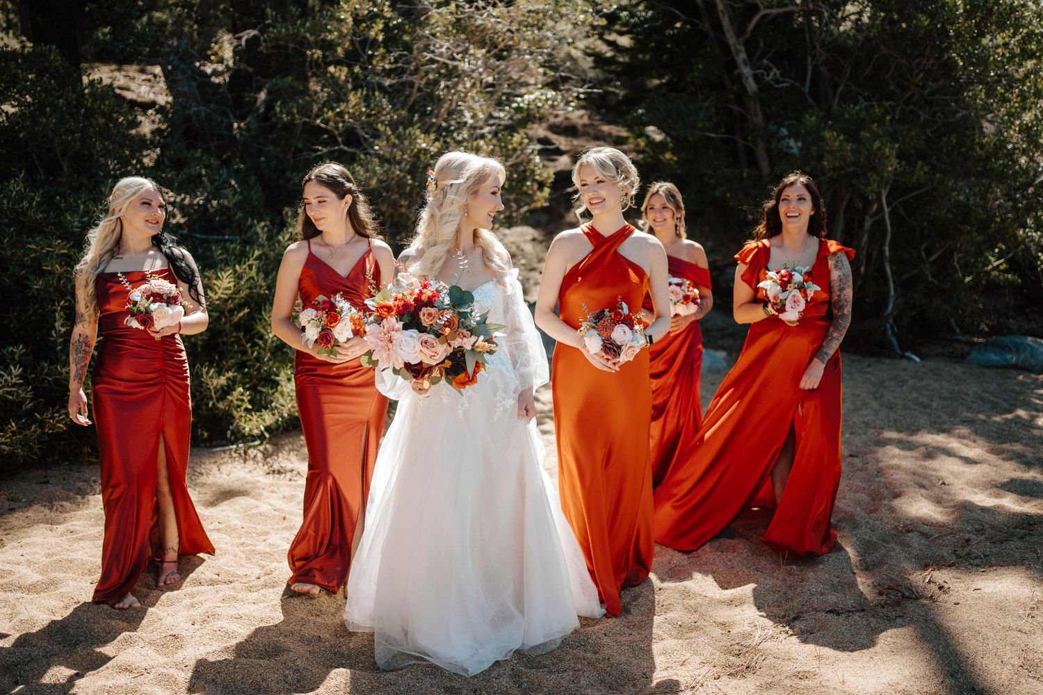 Bride walking with bridesmaids in orange dresses on a sandy outdoor path.