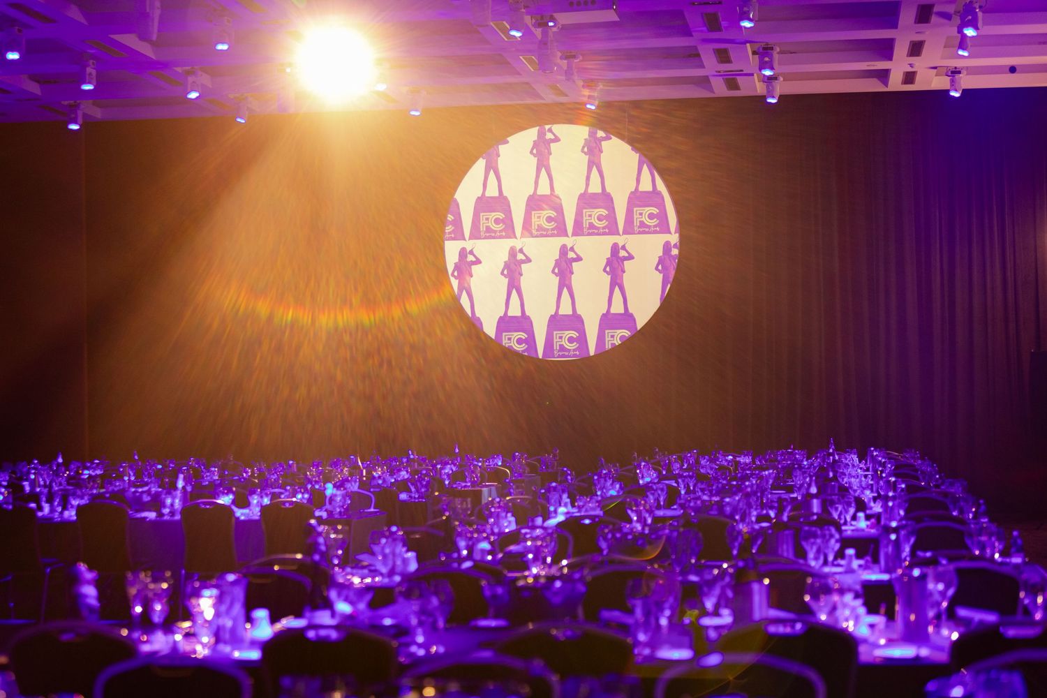 Blue and purple stage lighting illuminates a dark auditorium with rows of seats and geometric ceiling designs.