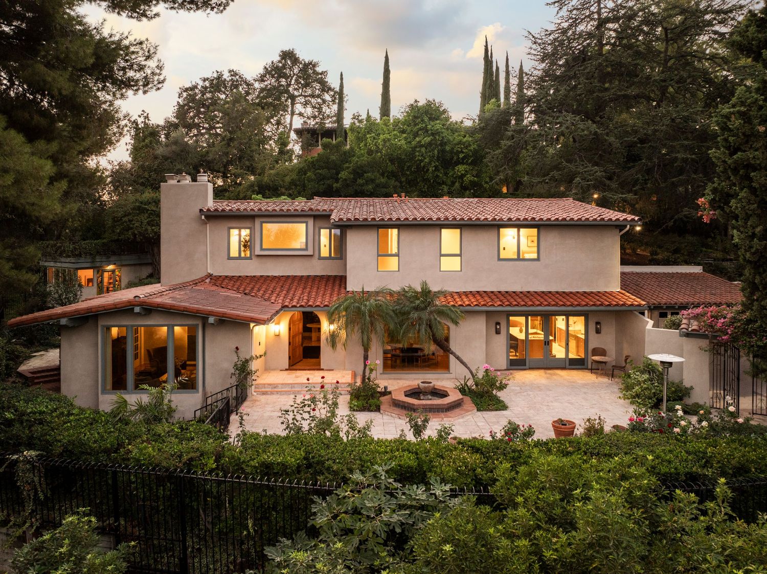 Mediterranean style luxury home with terracotta roof and glowing windows at dusk surrounded by lush landscaping.