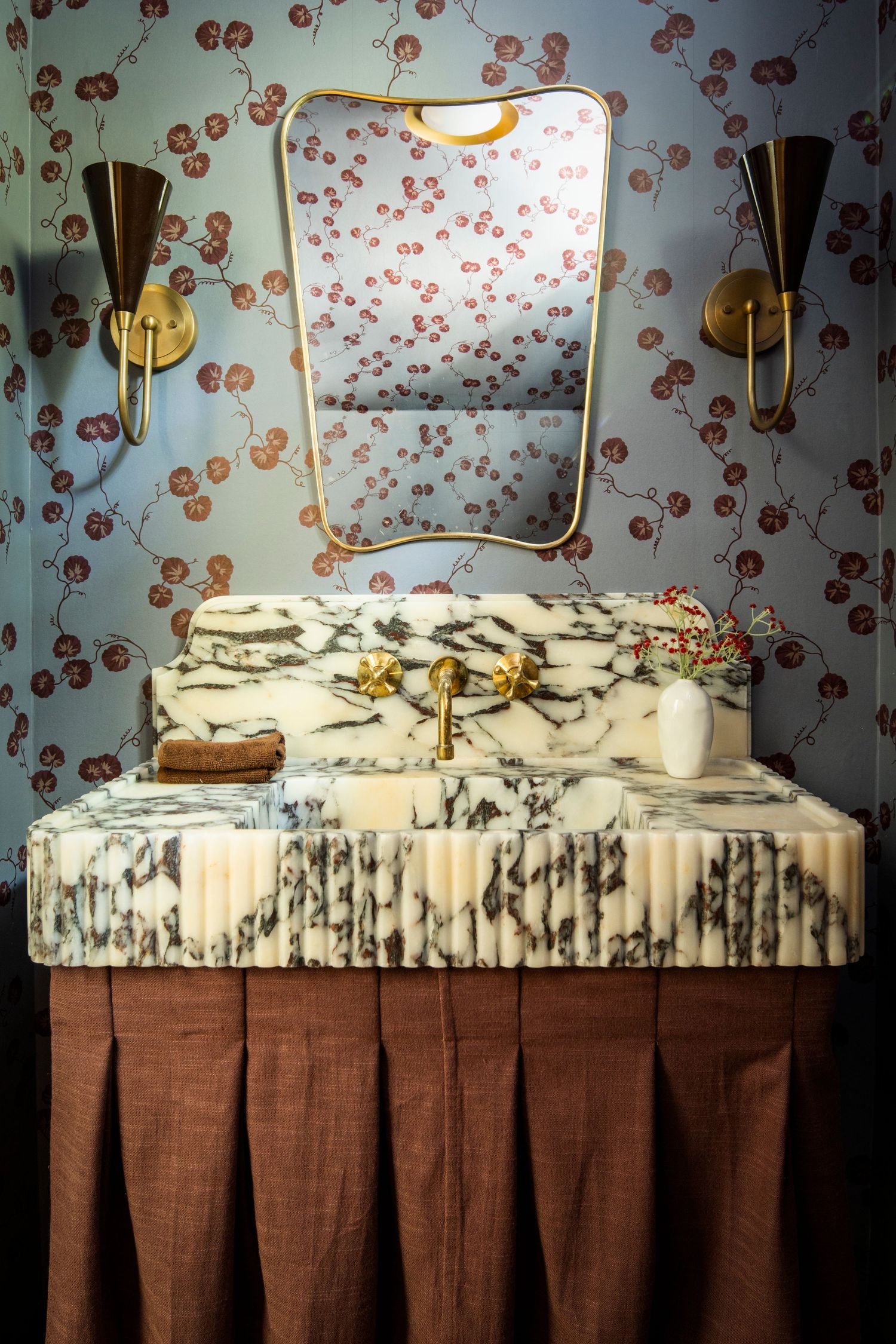 Vintage vanity with floral wallpaper backdrop and brass sconces features a marble top and brown pleated skirt.
