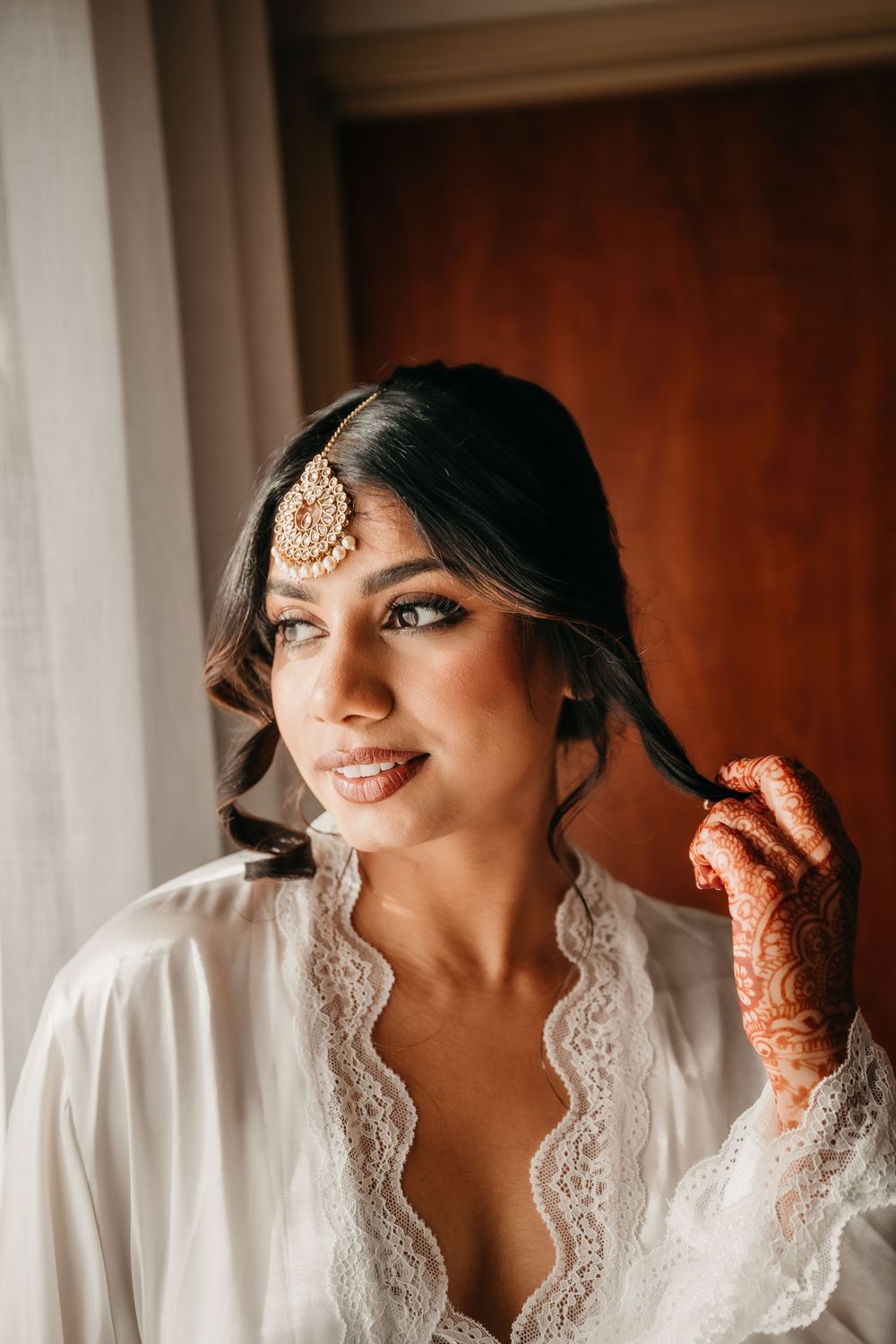 A bride in a white lace robe poses by a window during wedding preparations with traditional henna decorated hands.