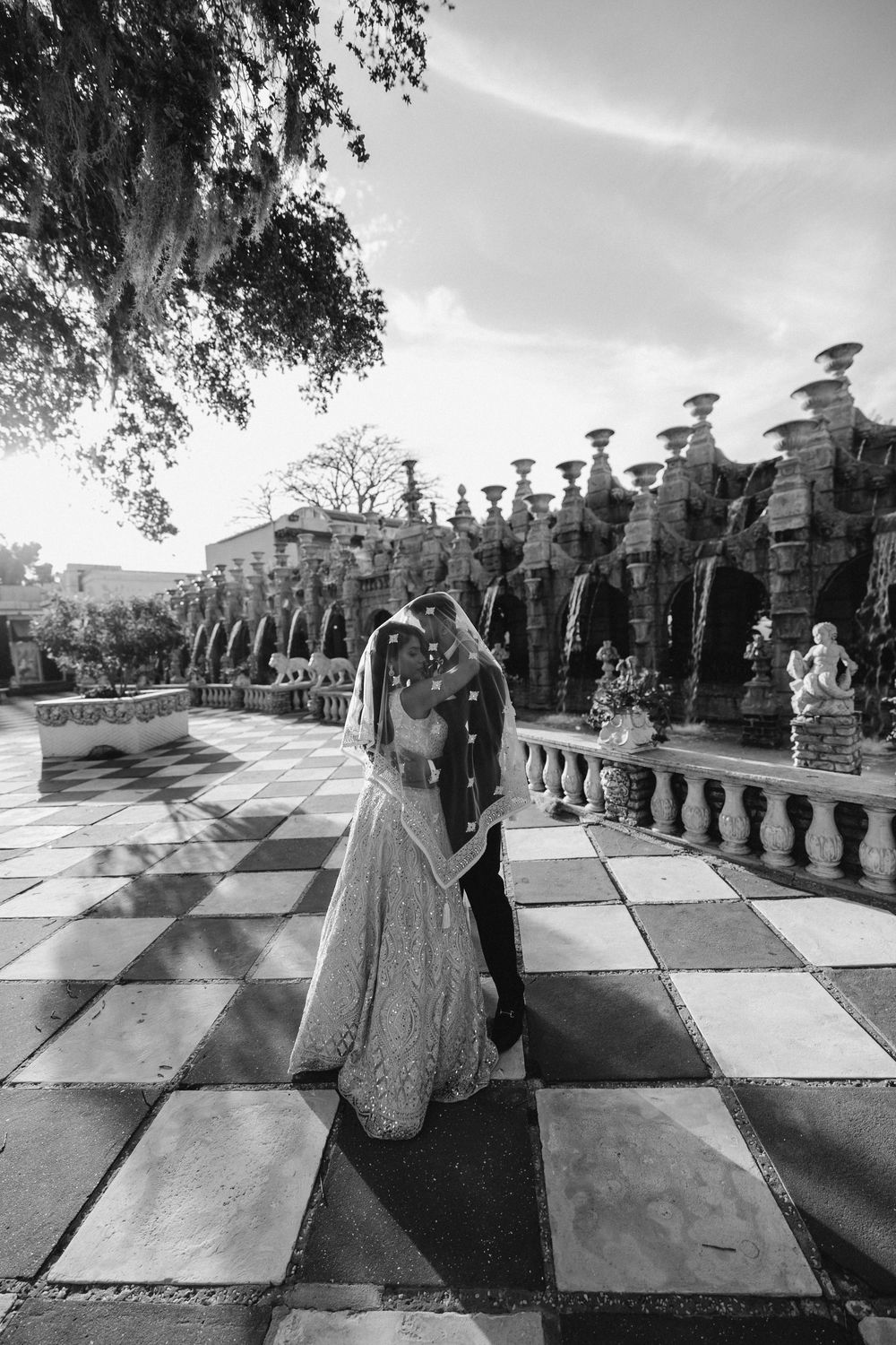 A romantic wedding couple embraces on a dramatic checkerboard terrace with ornate stone balustrades and columns.