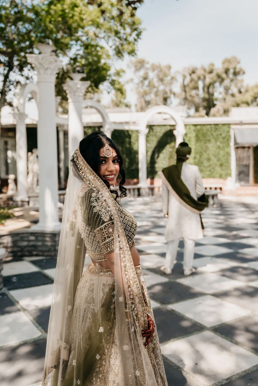 A bride in an ornate South Asian wedding dress poses gracefully on a checkered courtyard with white columns.