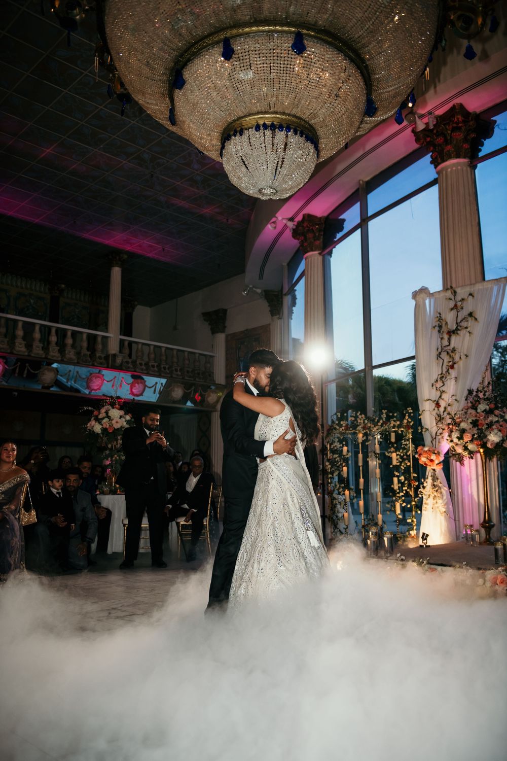A couple shares their first dance at an elegant wedding reception with fog effects and a crystal chandelier overhead.