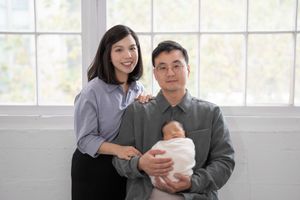 A happy family sits together on a couch near large windows while holding their newborn baby.
