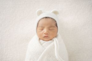 A sleeping newborn baby wrapped in white swaddling cloth lies peacefully on a soft white blanket.