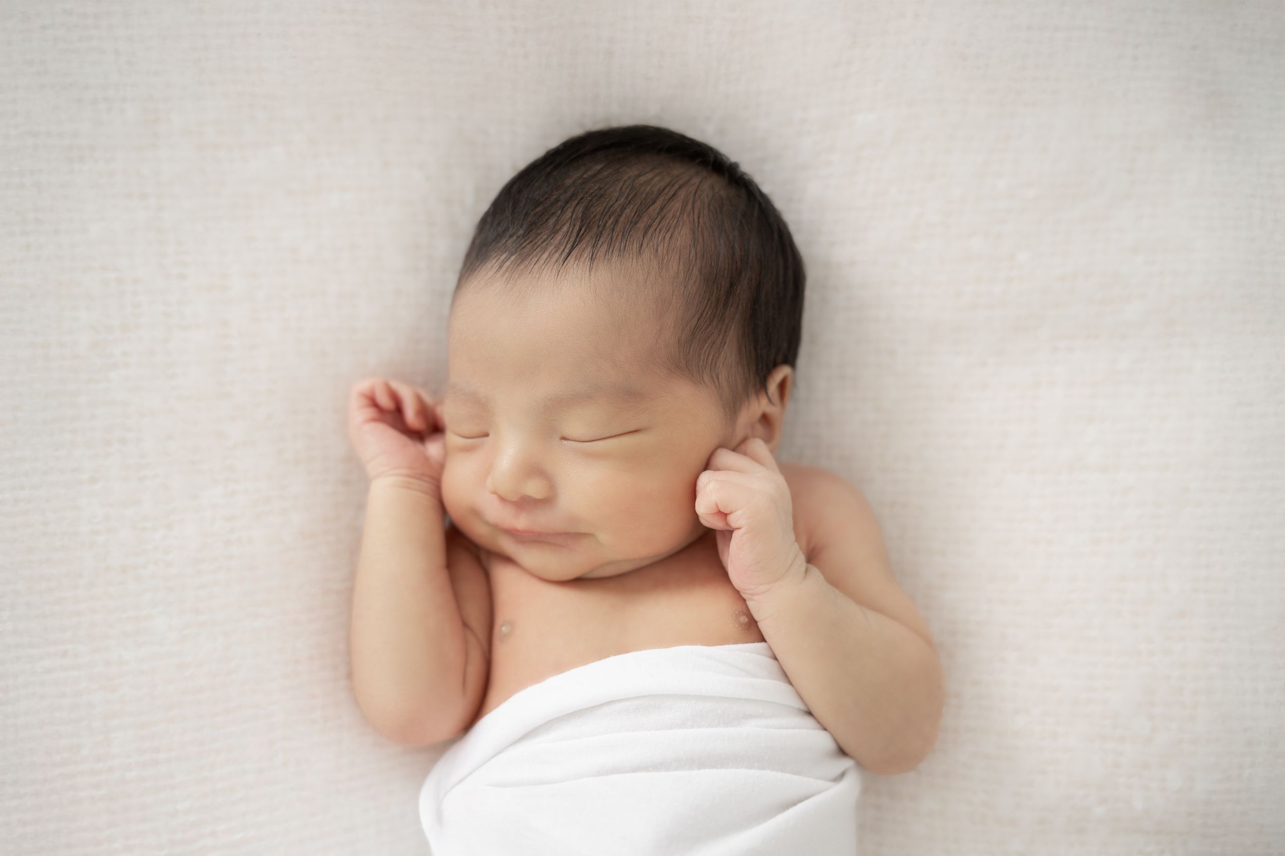 A sleeping newborn baby wrapped in white swaddle blankets rests peacefully during a professional photo session.