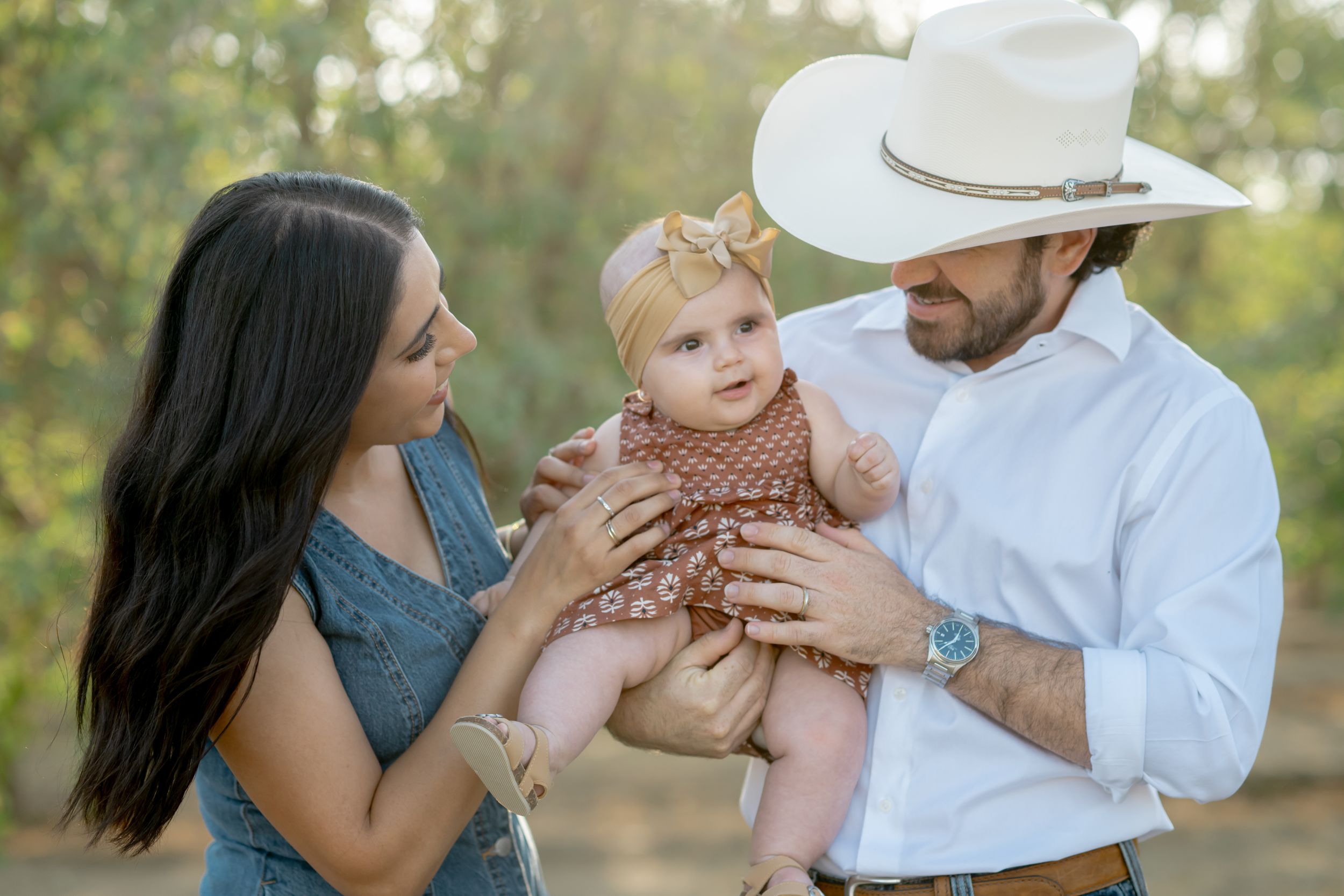 A family in casual attire shares a tender moment outdoors with their baby wearing a brown outfit.