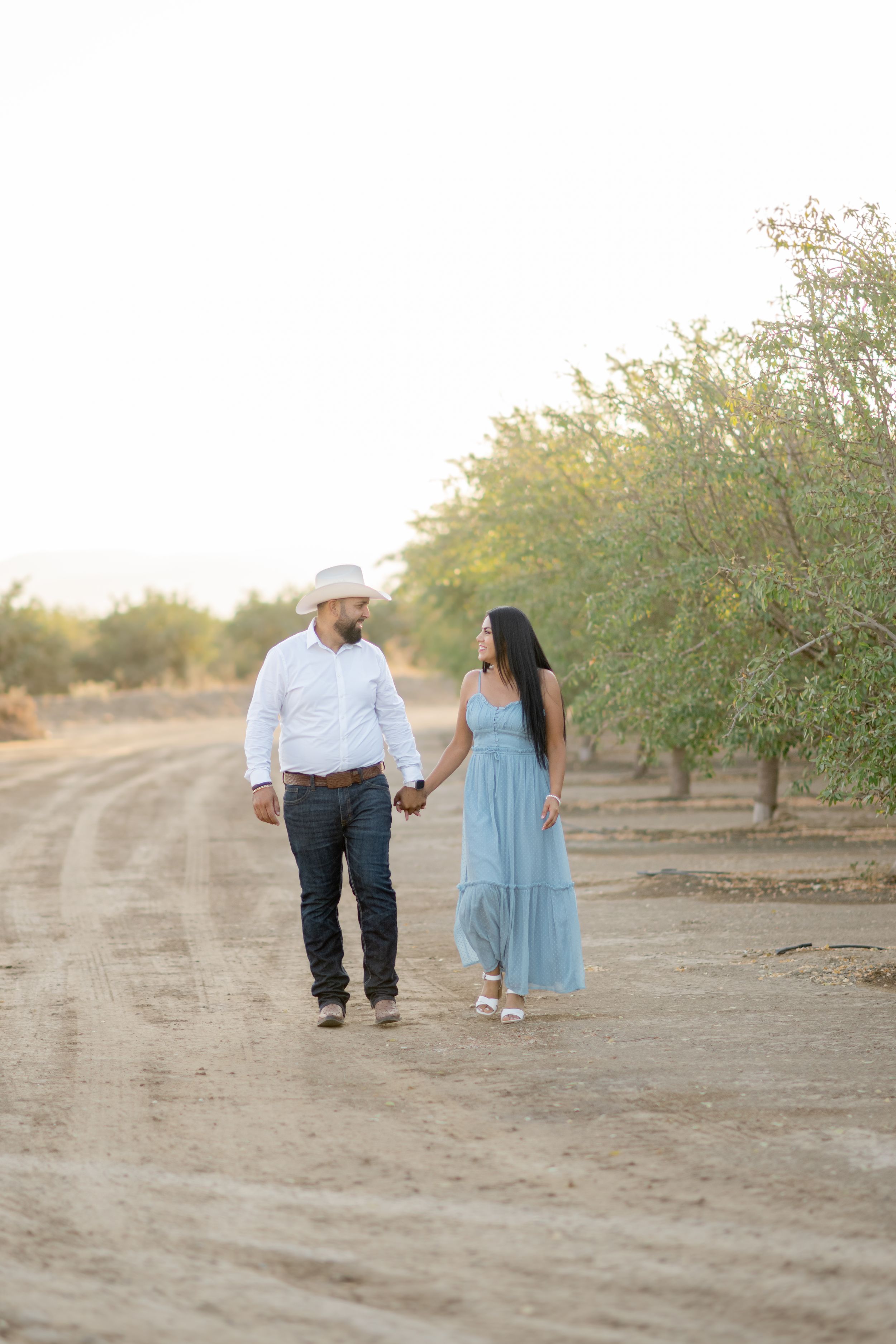 A couple in a light blue dress and white shirt walk together through a scenic almond orchard at sunset.