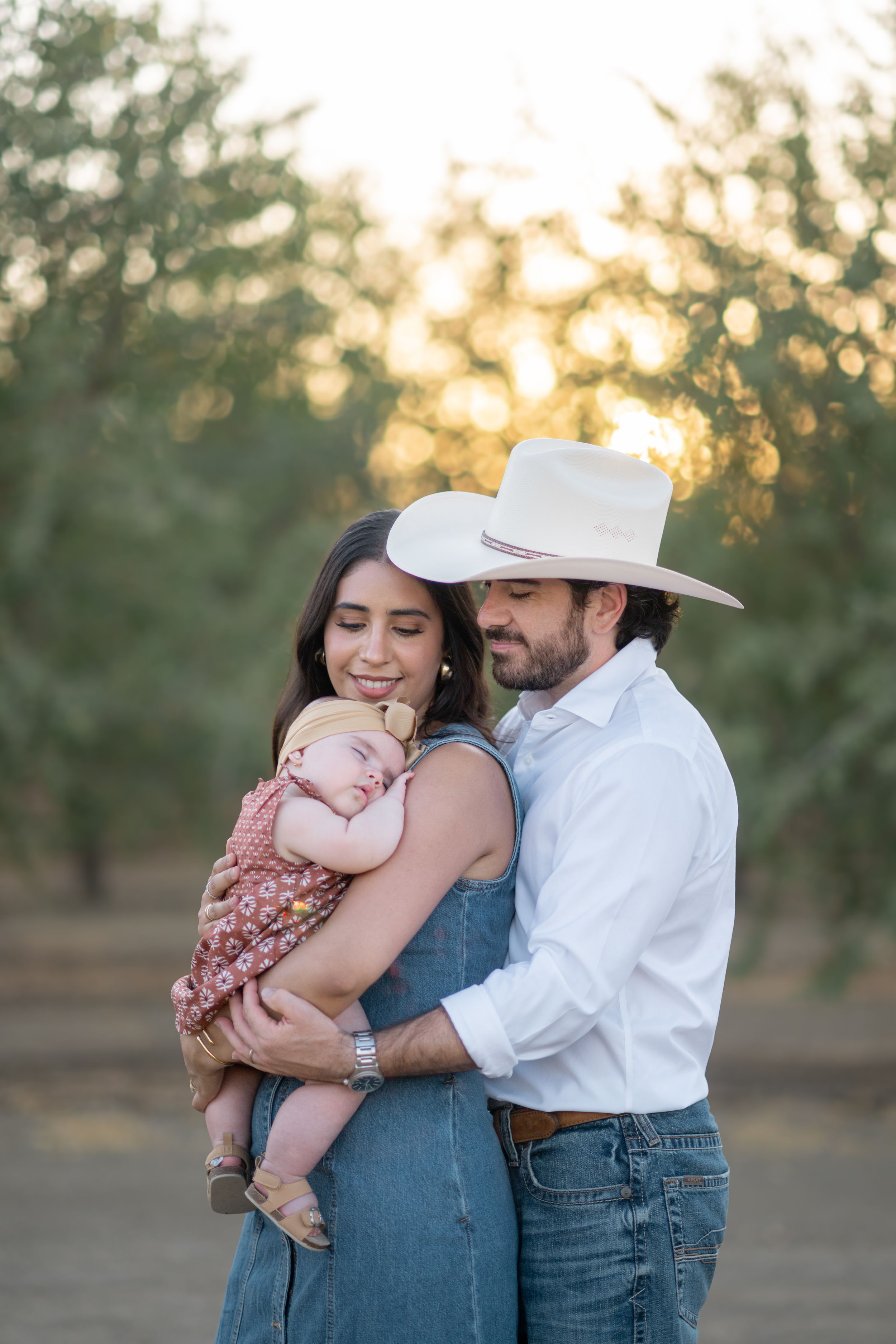 A couple in western attire shares tender moments with their baby outdoors during golden hour.
