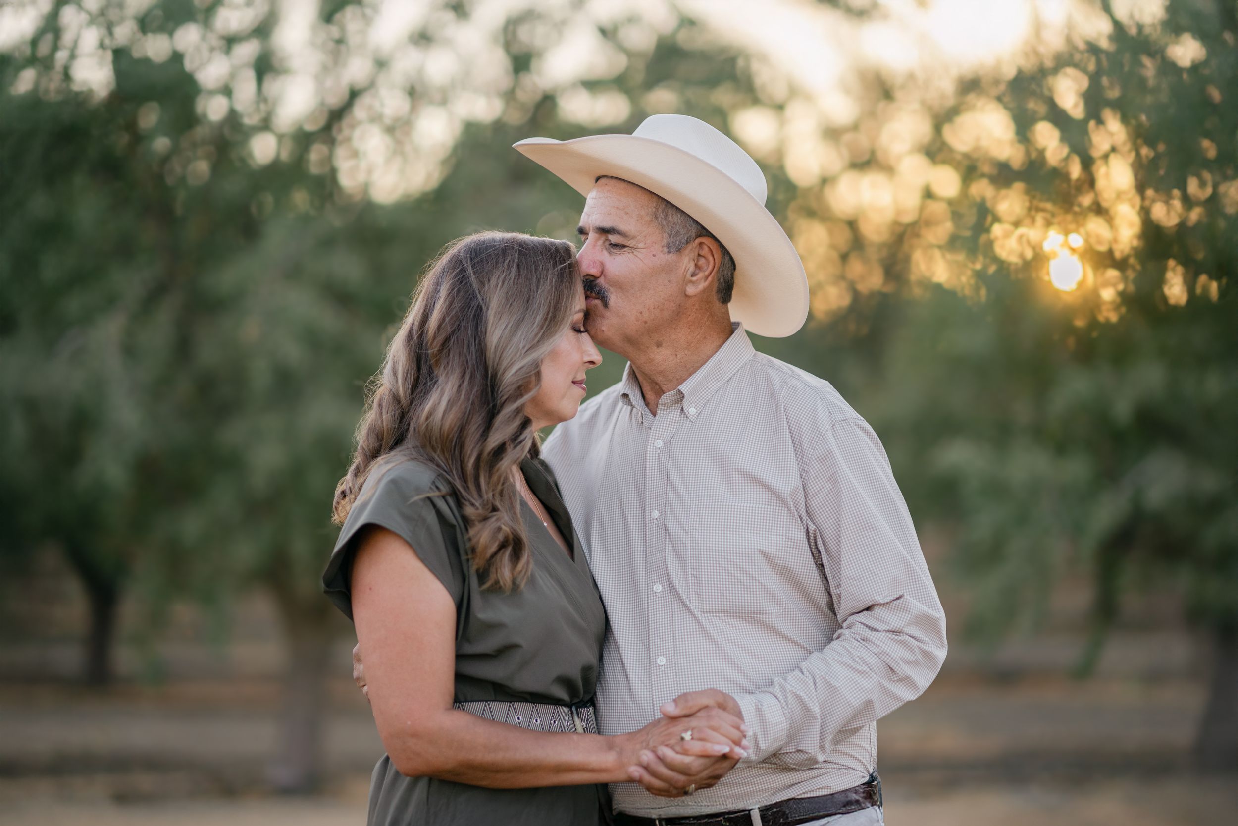 A couple in western attire shares romantic moments in an orchard during golden hour sunset.