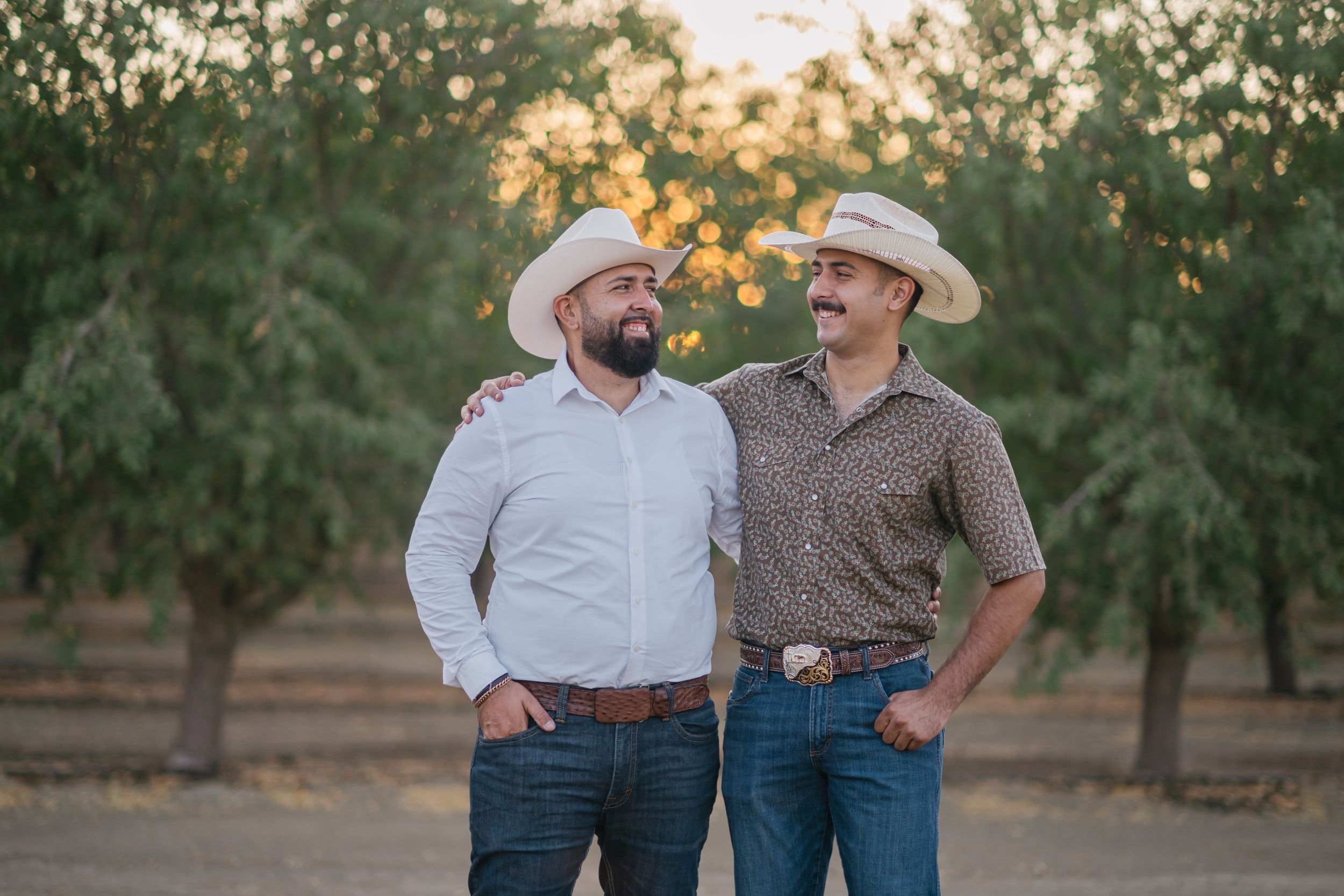 Two people wearing cowboy hats share a laugh while walking through an orchard at sunset.