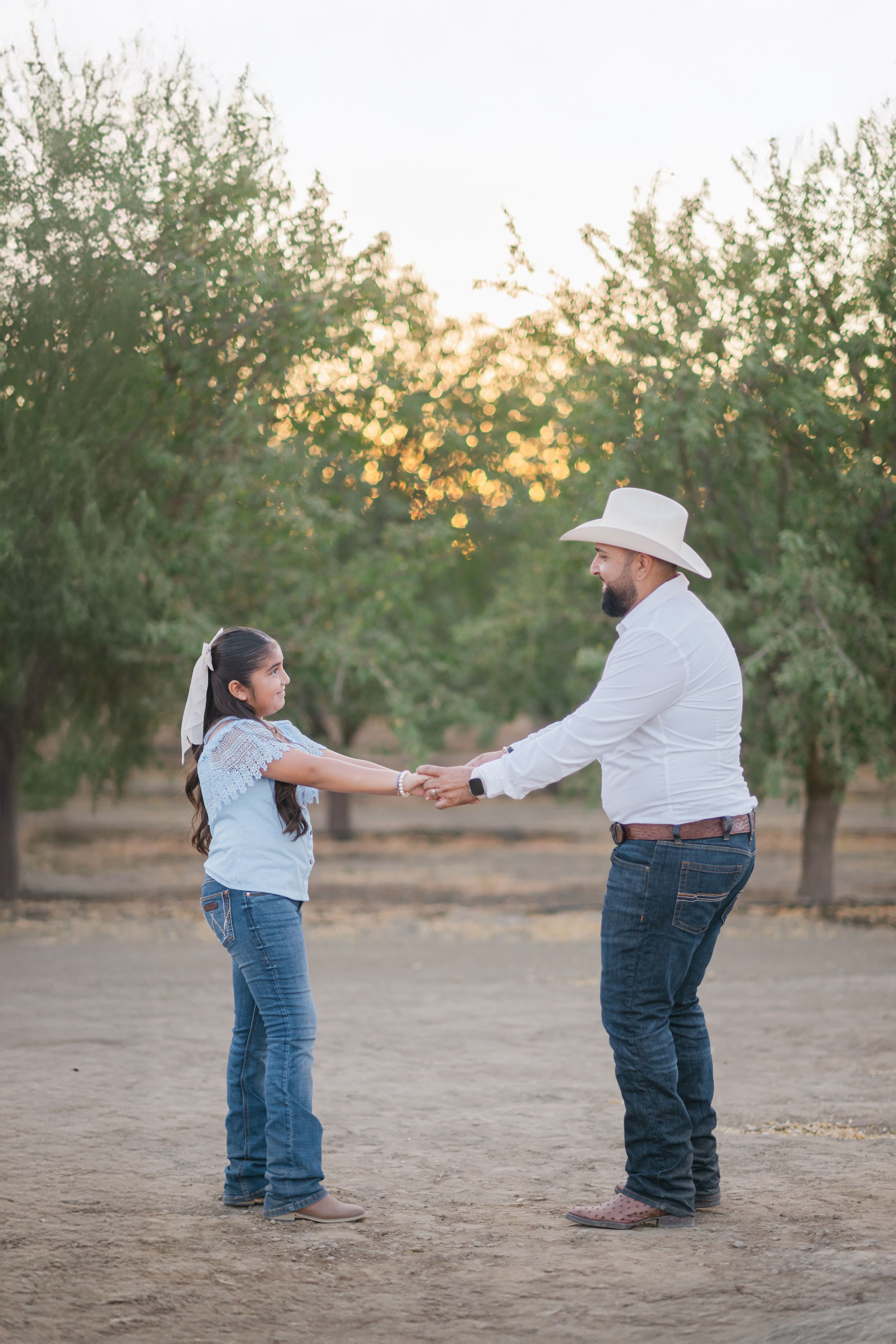 Two people in jeans and casual attire dance together in an orchard at sunset.