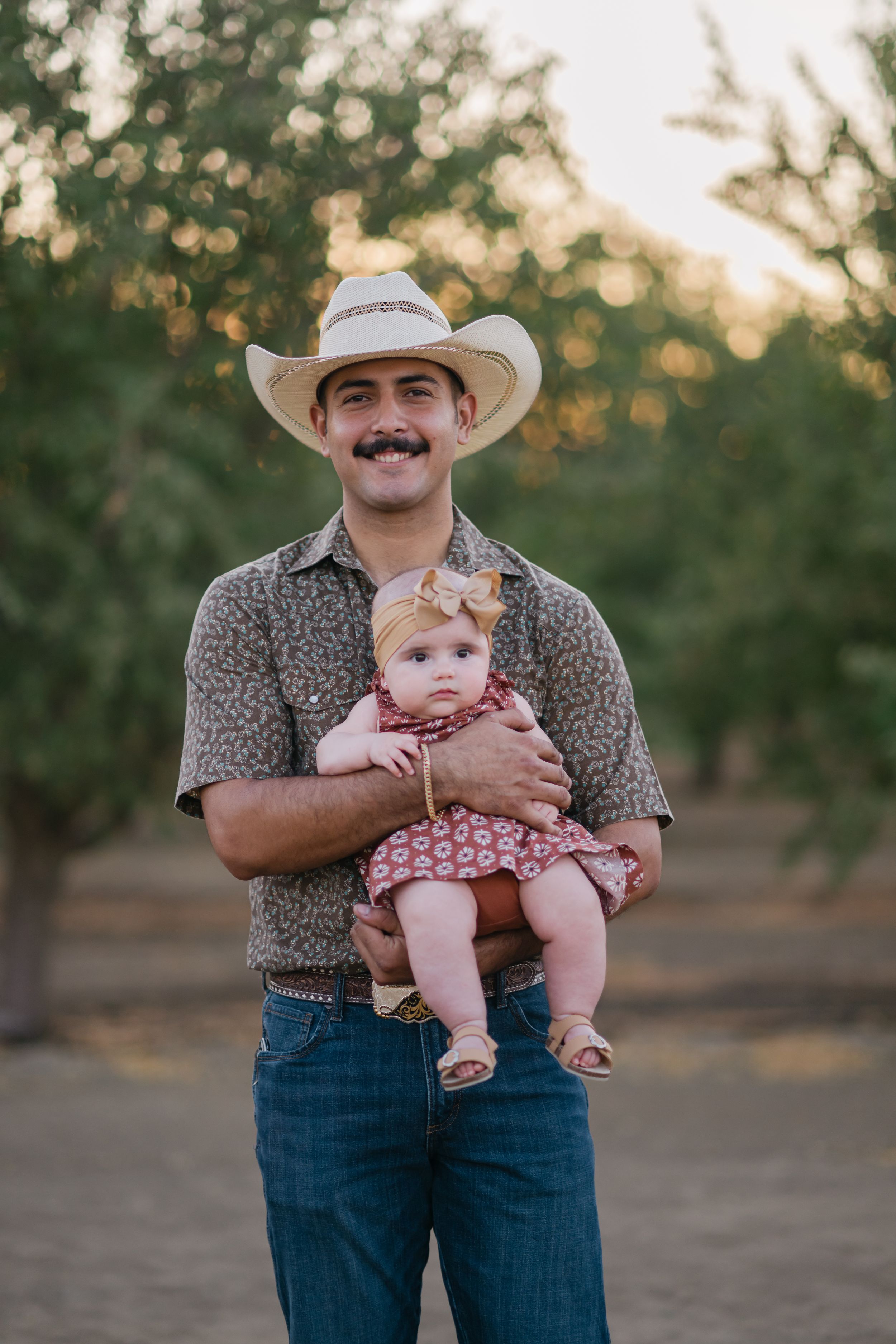 A person in a cowboy hat and patterned shirt holds a small baby in a pink dress outdoors at sunset.
