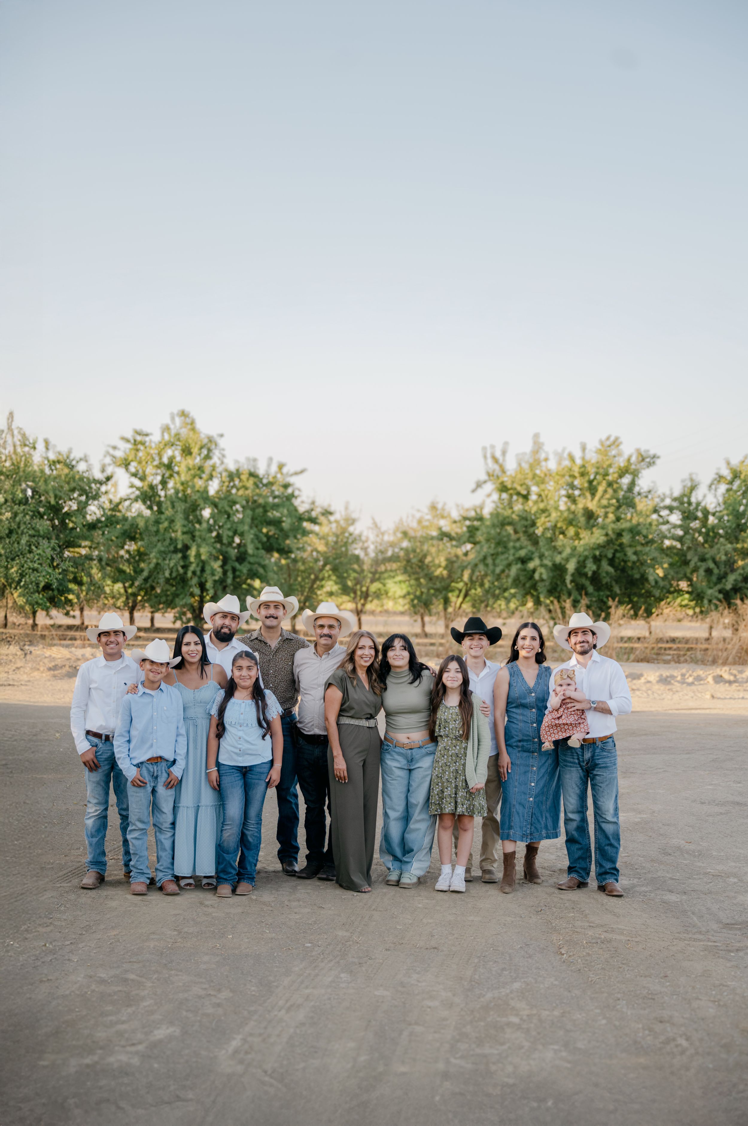 A group of people in casual denim and neutral clothing stand together in a line outdoors against a rural backdrop.