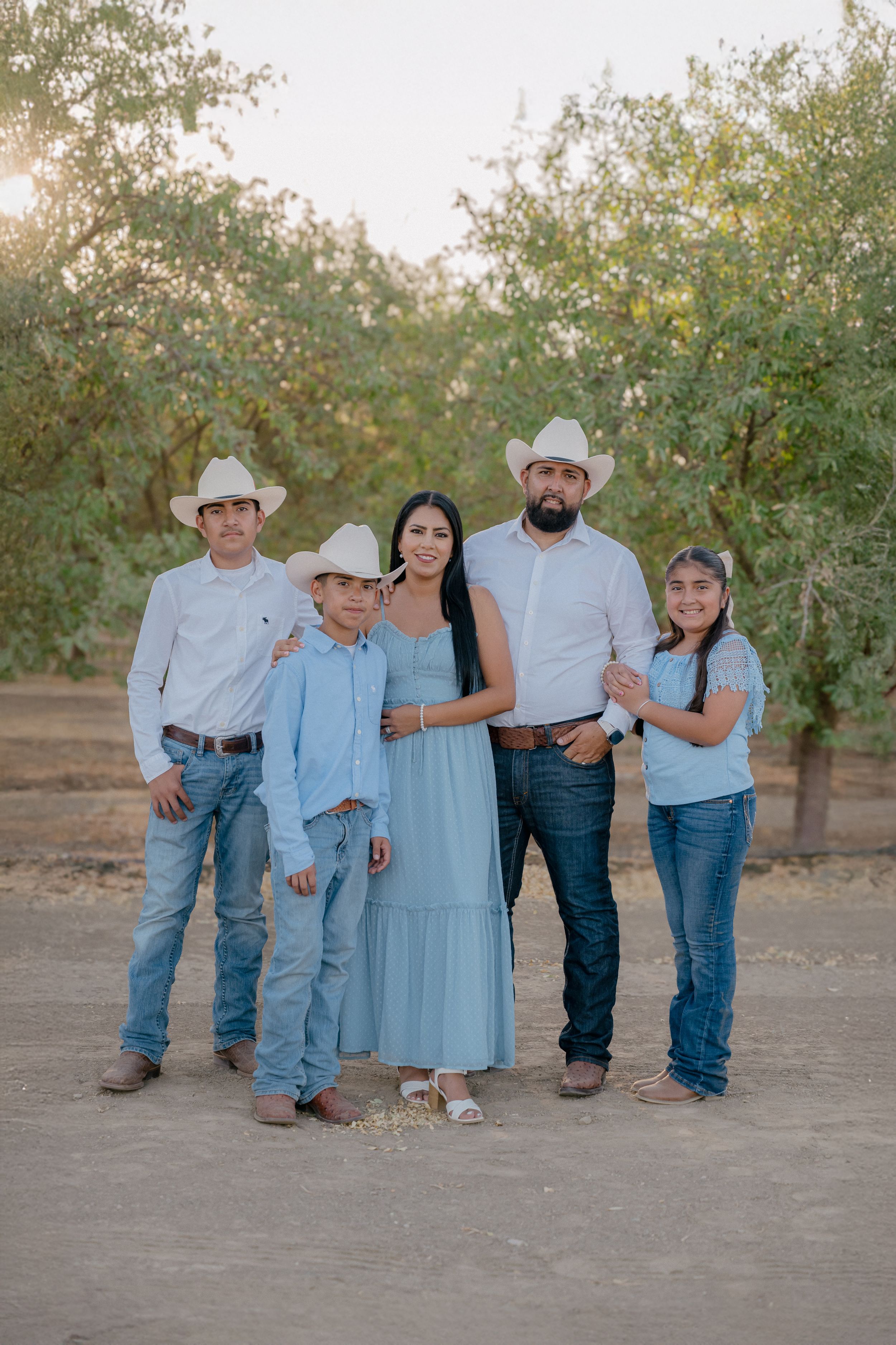A family in coordinating blue and white outfits poses together on the ground at sunset in an outdoor setting.