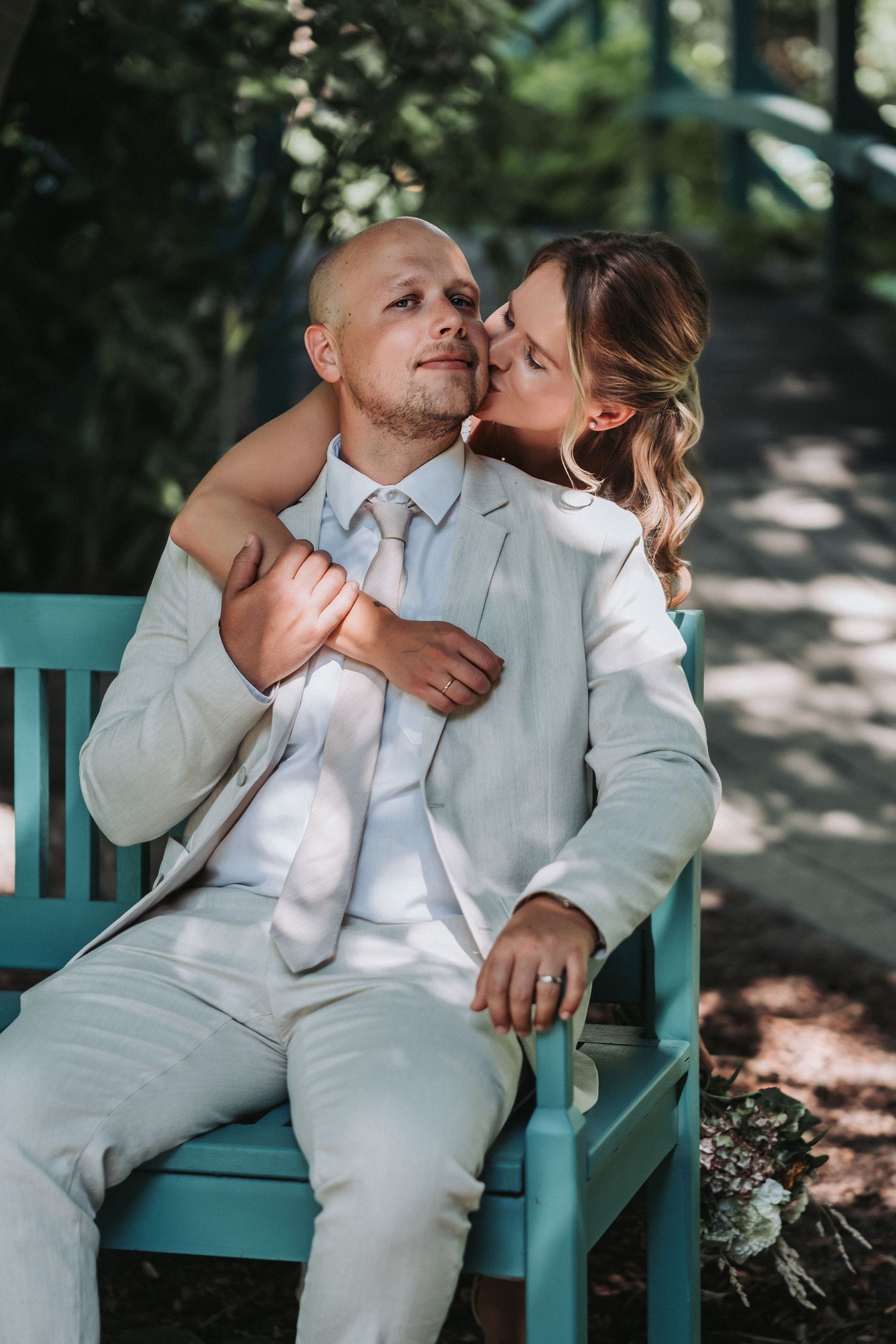 A couple in white outfits shares tender romantic moments while sitting together on a turquoise bench in a garden setting.
