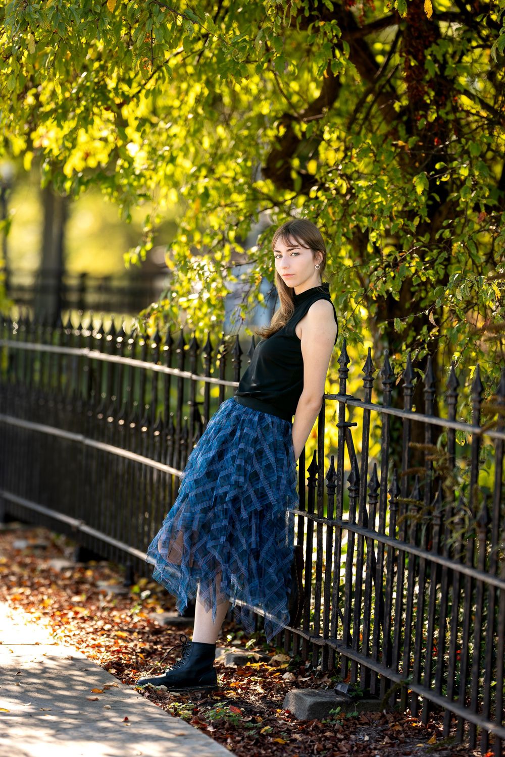 Elegant woman in blue patterned midi skirt and black top standing by wrought iron park fence.