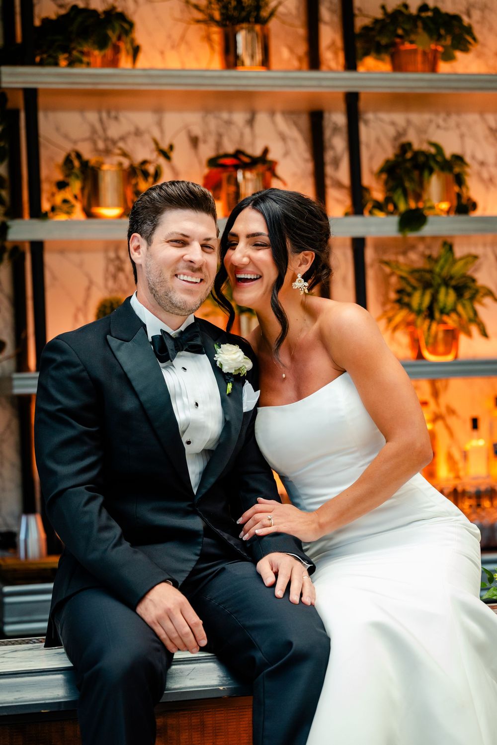 A joyful couple shares a candid moment at their wedding reception with potted plants and warm lighting in the background.