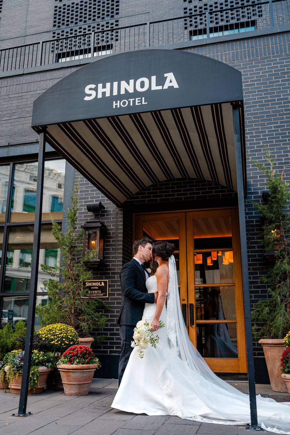 A wedding couple stands outside the entrance of the Shinola Hotel with a flowing white wedding dress and veil.
