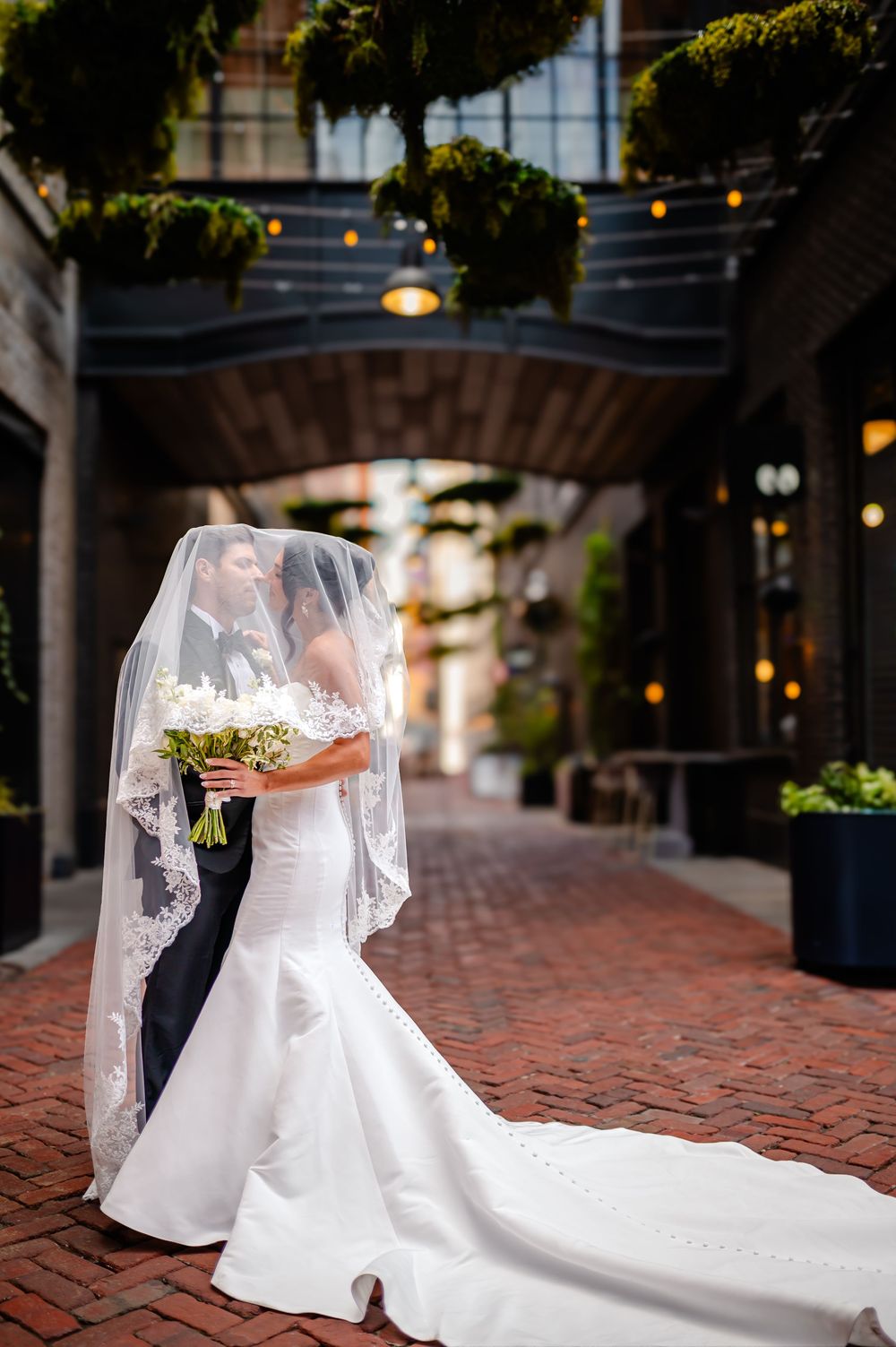 A wedding couple poses along a brick-paved walkway with string lights and a flowing white dress train.