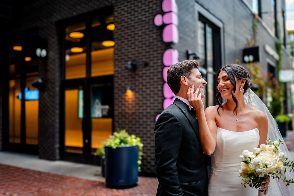 Newlywed couple shares a romantic moment together outside a brick building with pink lighting.