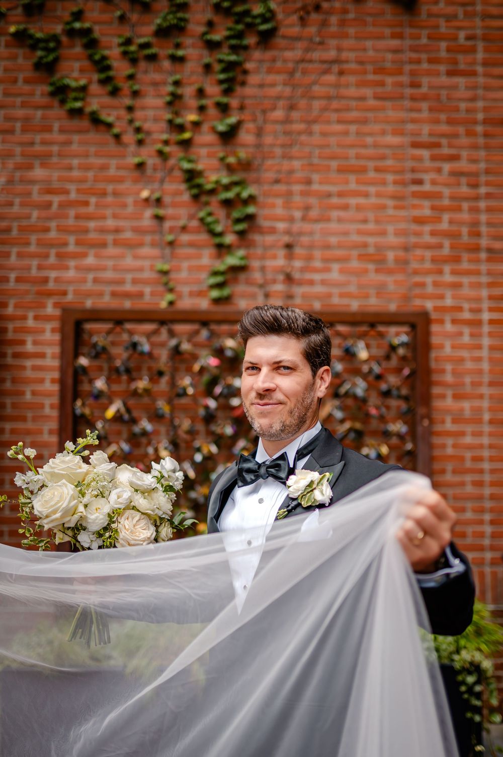 Wedding party members adjust a long flowing veil against a brick wall backdrop with climbing ivy.