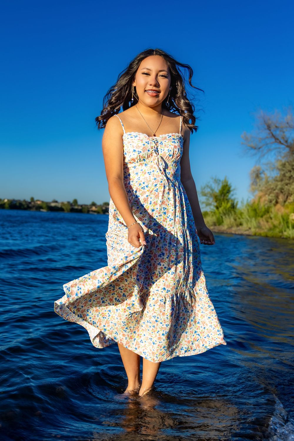 Person in floral sundress stands in shallow water against blue sky and lake backdrop as dress flows in breeze.