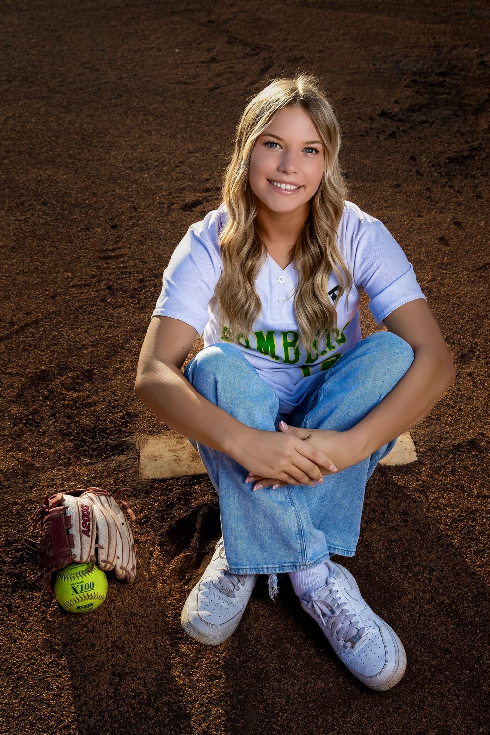 Softball player in blue and white uniform sits cross-legged on dirt field with glove and ball nearby.