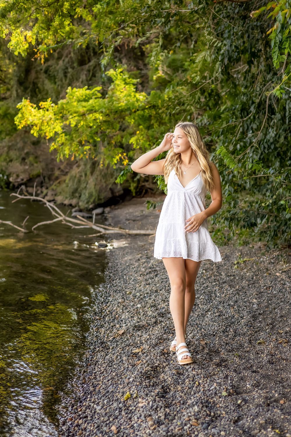 Someone in a white dress walks along a rocky riverbank surrounded by lush green trees and sunlit foliage.