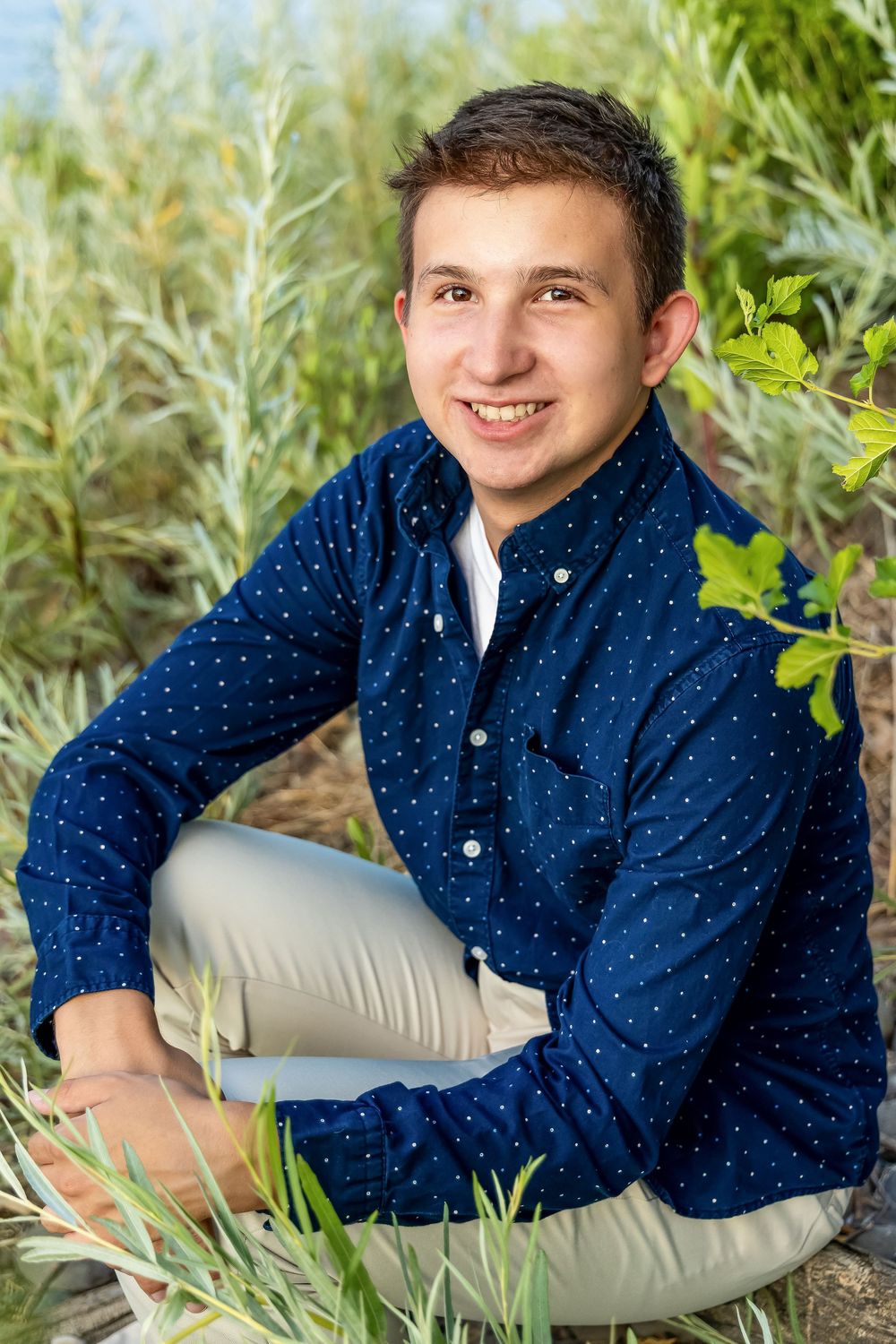 Outdoor portrait of someone wearing a navy polka dot button-down shirt and khaki pants sitting among green foliage.