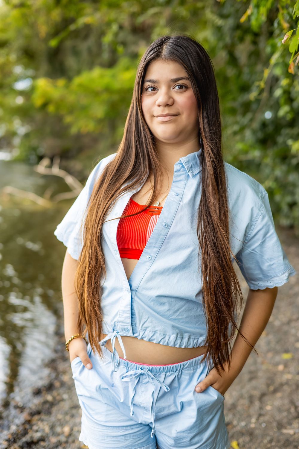 A person in a light blue button-up shirt and red top poses outdoors against a natural background with trees and water.