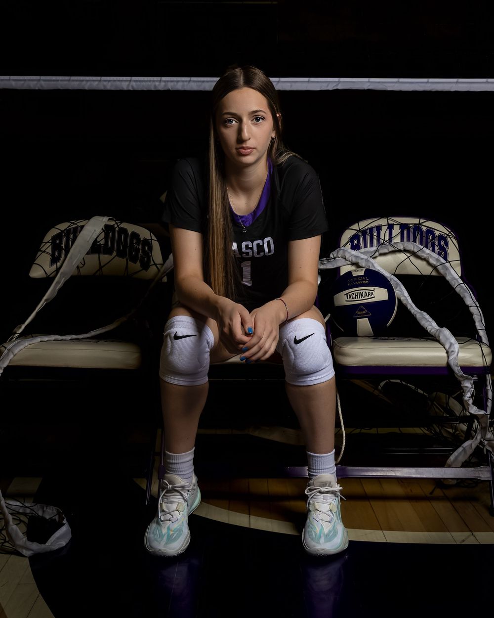Athlete in black t-shirt and knee pads sits on bench between lacrosse sticks in dramatic dark lighting.