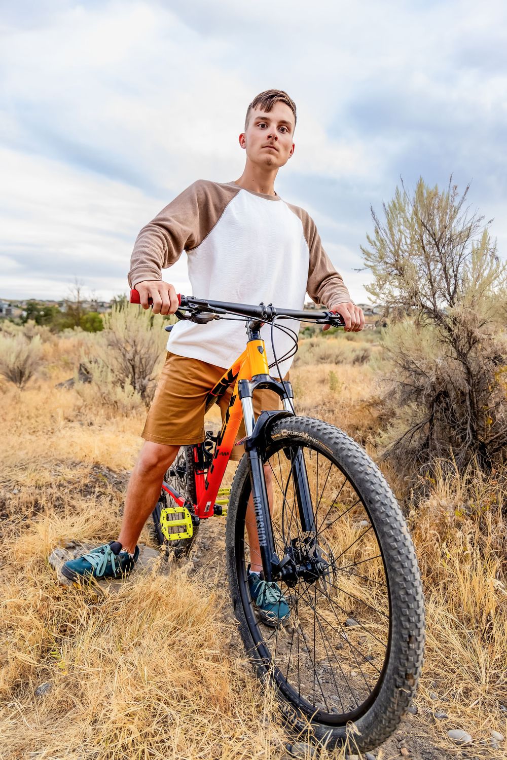 Mountain biker with orange bike stands on dry terrain against cloudy sky in desert landscape.