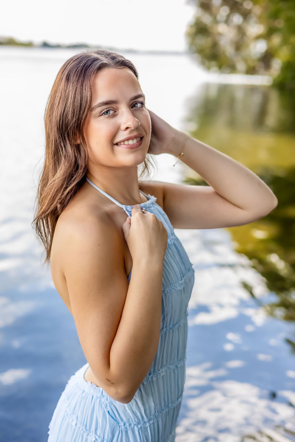 Woman in light blue sundress poses outdoors near water on sunny day with natural lighting and blurred background.