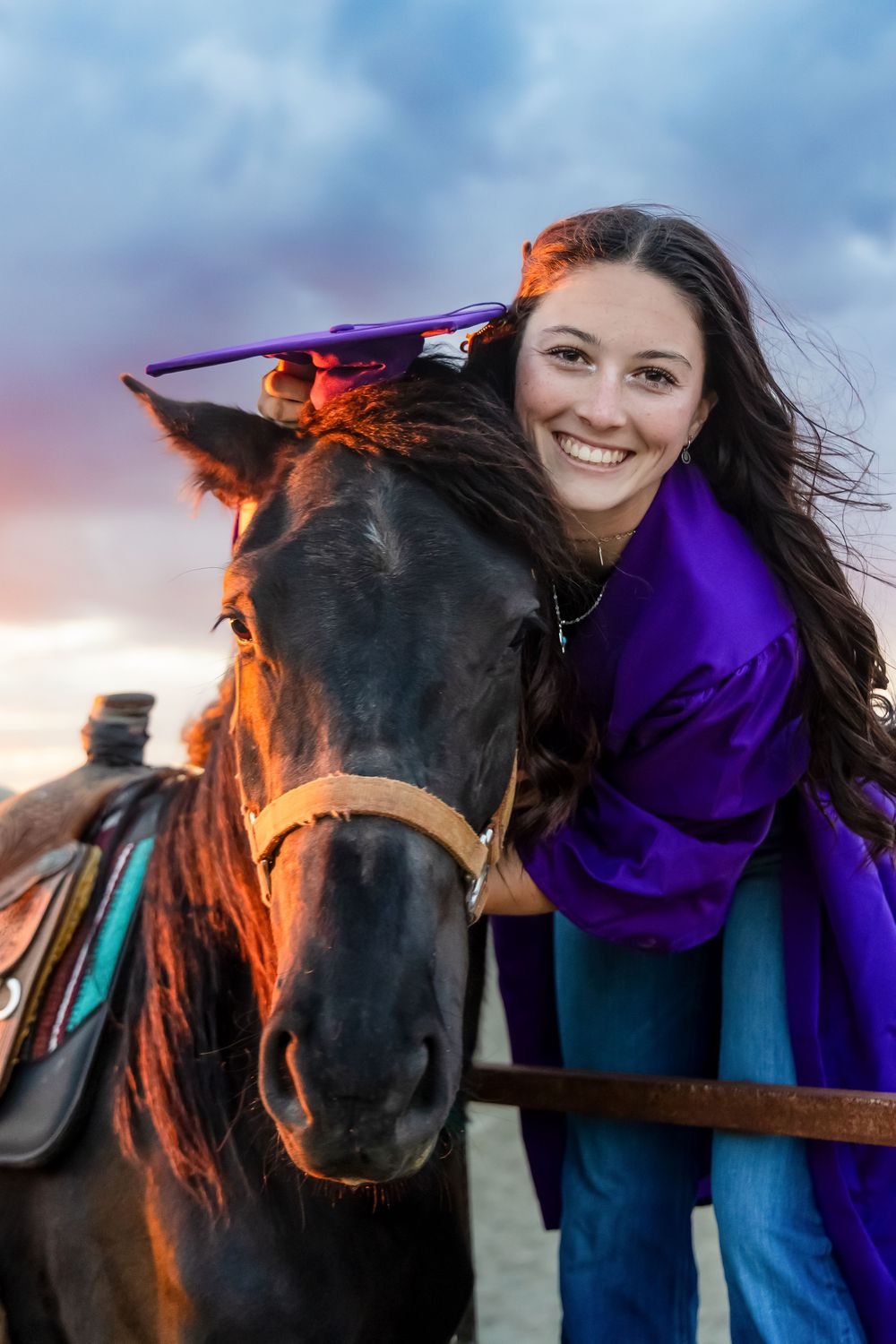 A person in a purple sweater shares tender moments with a black horse against a cloudy sky at sunset.
