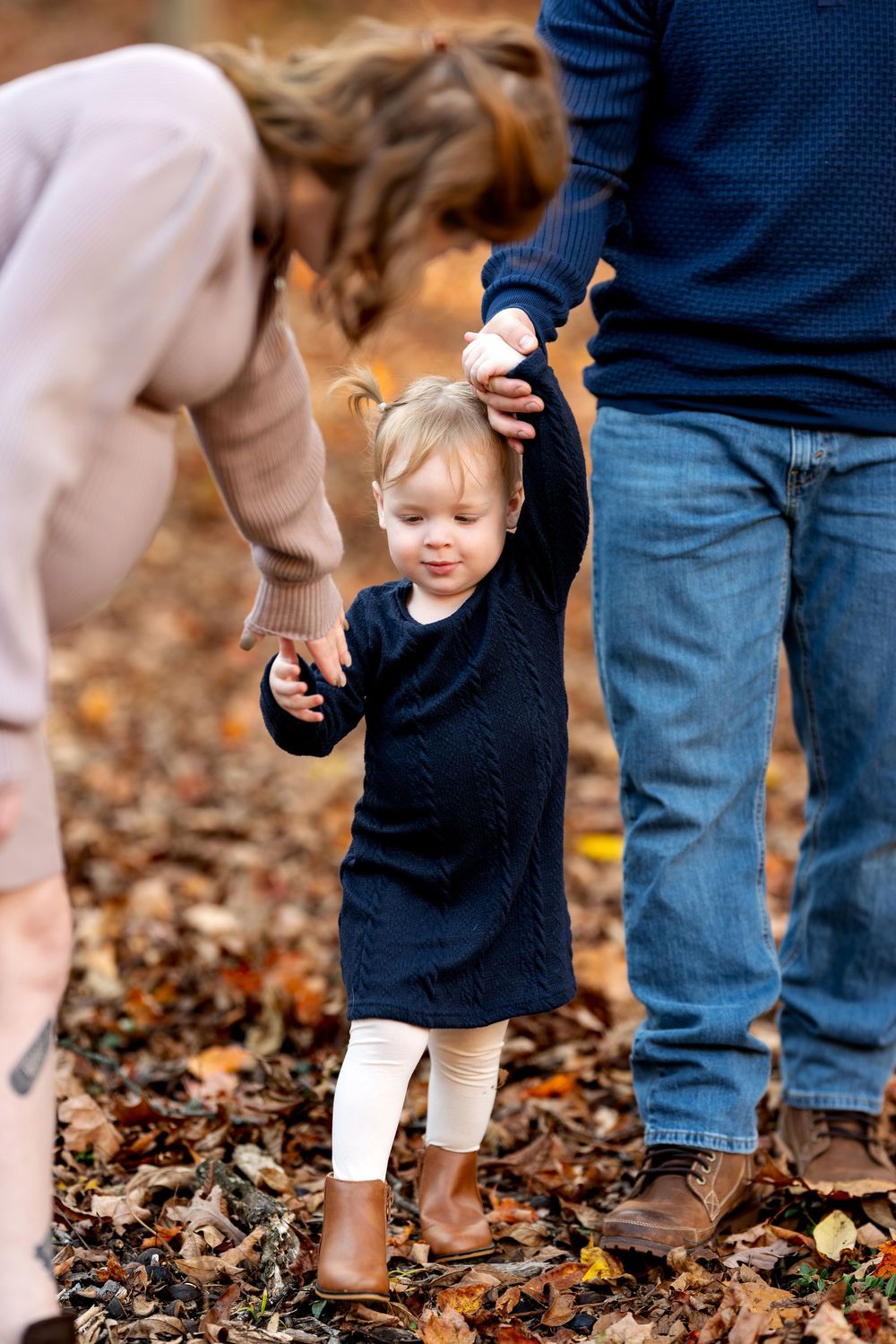 Parents holding toddler girl's hands while walking through autumn leaf-covered ground.